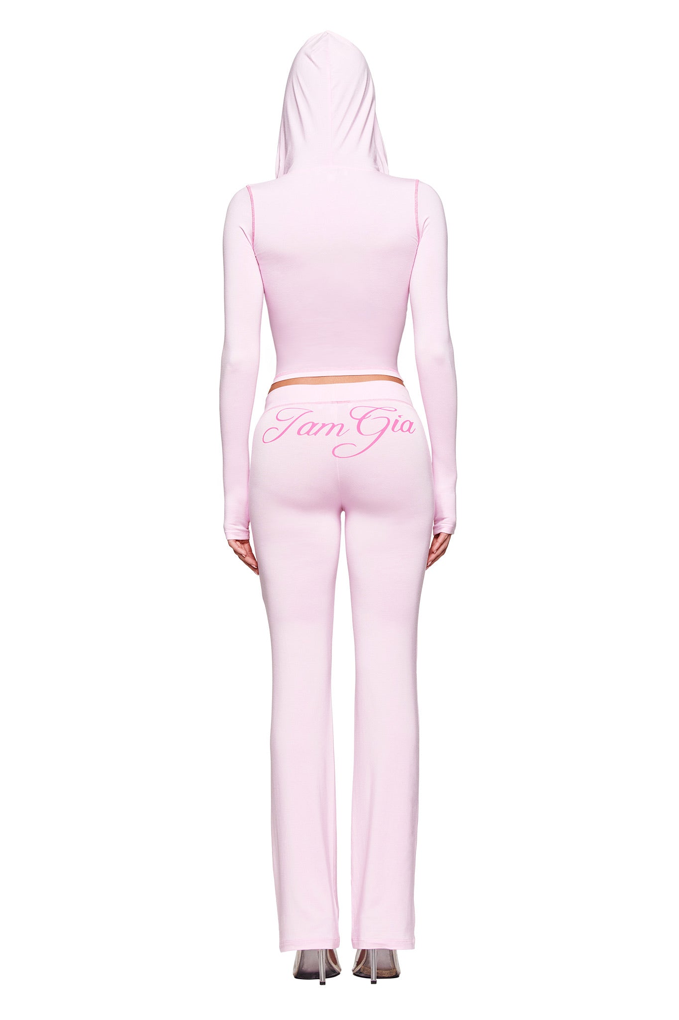 A person is seen from behind wearing the I.AM.GIA BLARE HOODIE in pastel pink, styled as a cropped bodycon top with matching high-waisted pants featuring "I am Gia" in pink cursive across the back.