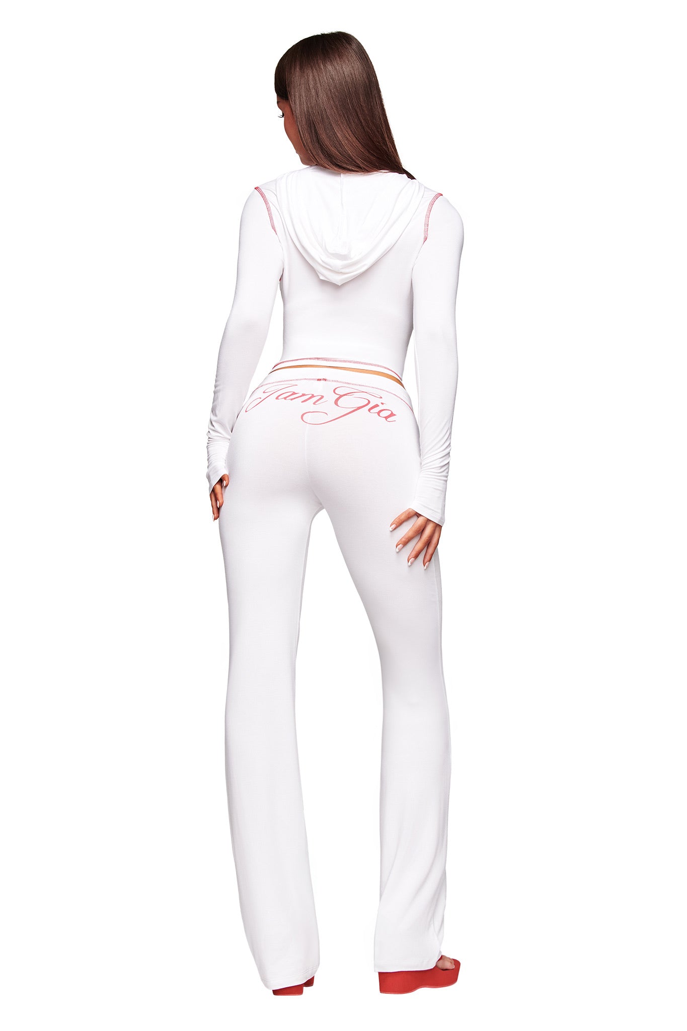 A woman with long brown hair wears I.AM.GIA’s fitted white hoodie and BLARE TRACKPANT - WHITE, featuring “Gia Gen Gia” in pink script on the low-rise back, styled with red platform shoes for a bold, signature look.