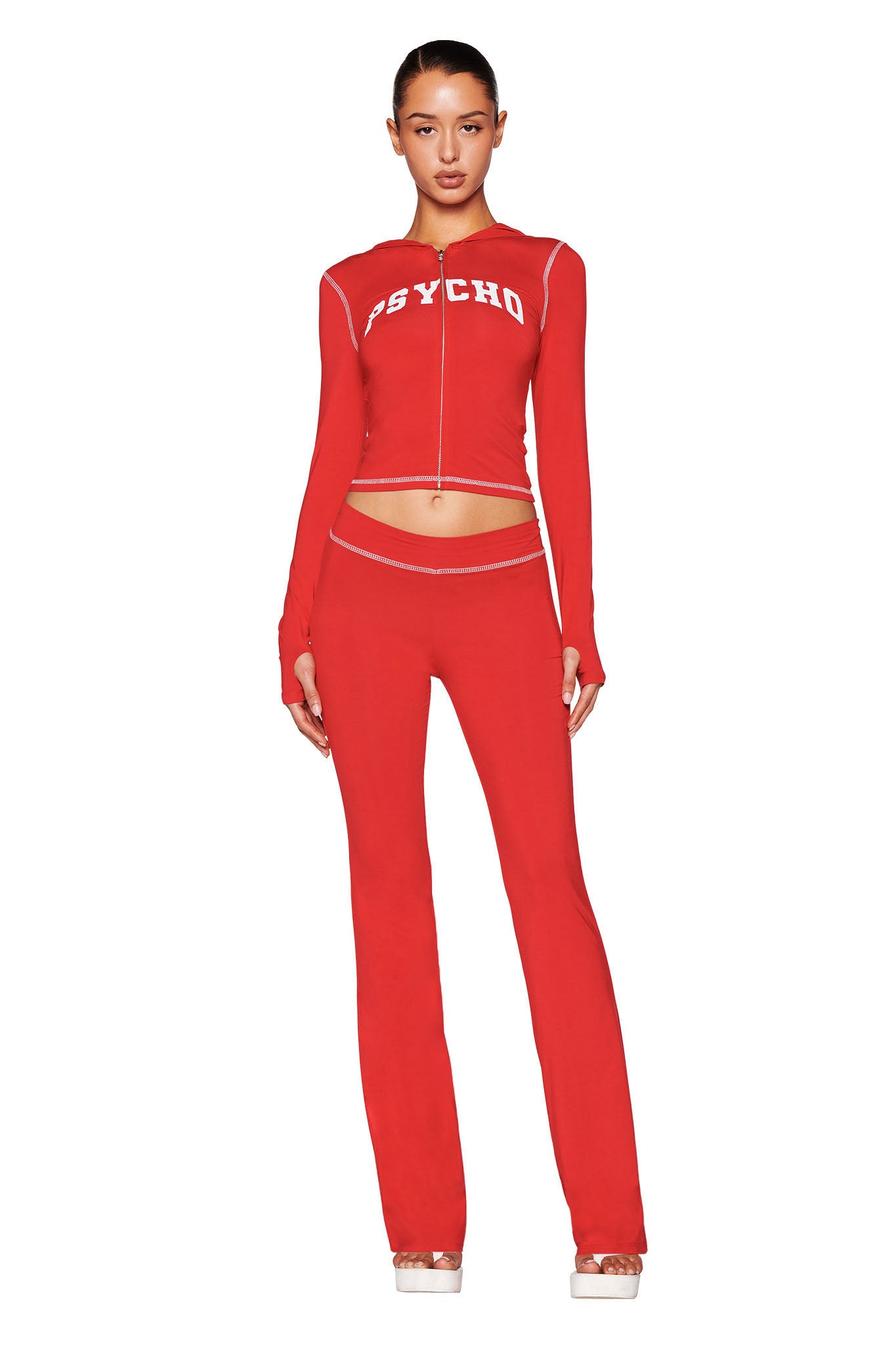 A woman faces forward in a fitted red I.AM.GIA BLARE TRACKPANT - RED : PSYCHO : SALSA set with long sleeves, midriff cutout, "PSYCHO" printed in white on the top, and platform shoes.