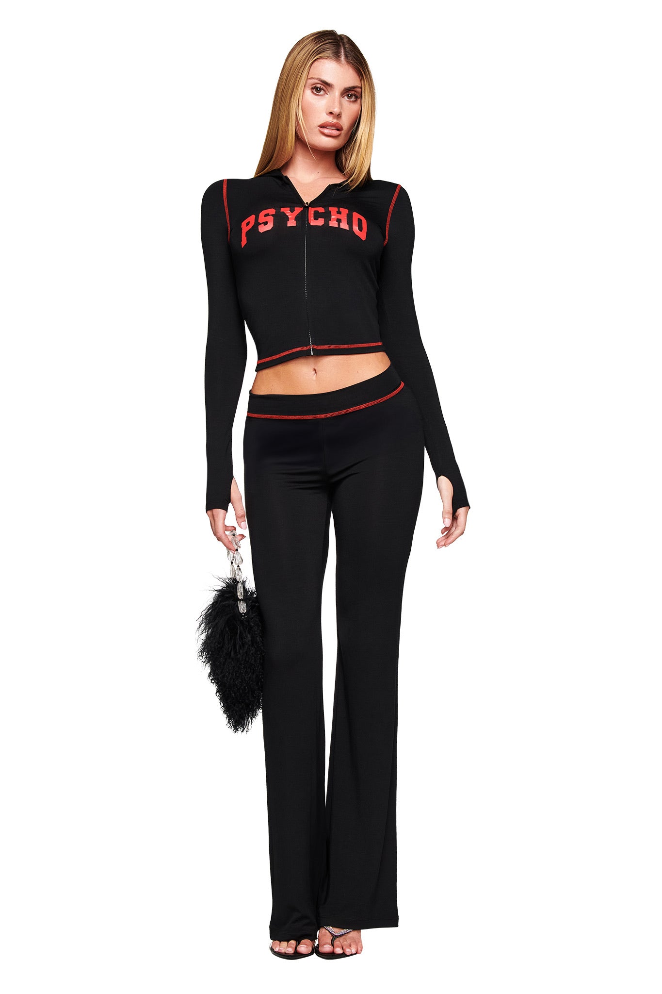 A woman faces forward in I.AM.GIA’s BLARE TRACKPANT - BLACK PSYCHO, a black two-piece with bodycon fit and red trim; the top features bold red "PSYCHO" lettering. She holds a black furry handbag and wears open-toed black shoes on a white background.