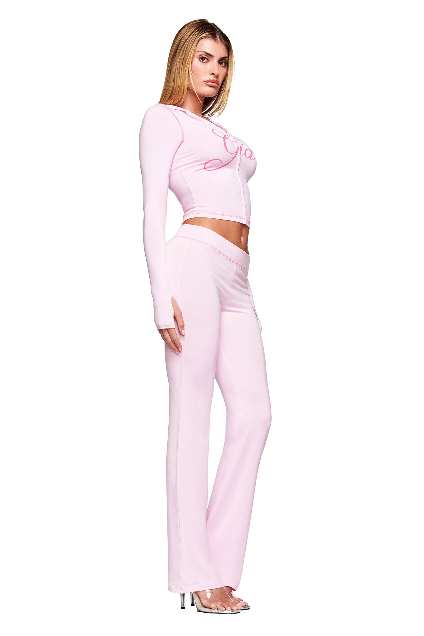 A woman in profile models the I.AM.GIA BLARE HOODIE in pastel pink, styled as a long-sleeve crop top with matching flared pants and high-heeled sandals, set against a white background.