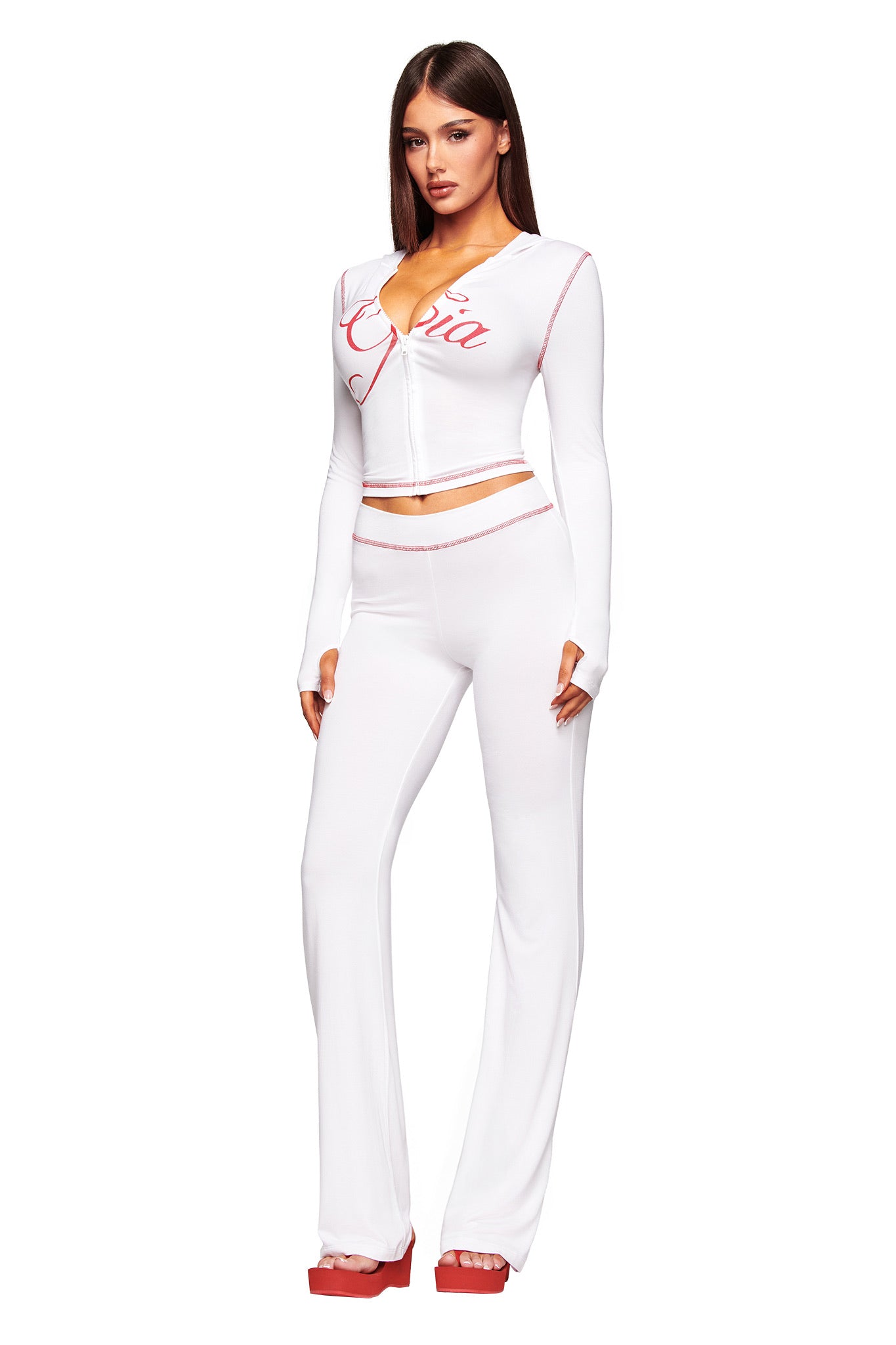 A woman stands against a white background in I.AM.GIA’s fitted BLARE TRACKPANT in white, featuring a low-rise waist, red trim accents, and paired with a matching long-sleeve top with red script on the chest and red platform shoes.