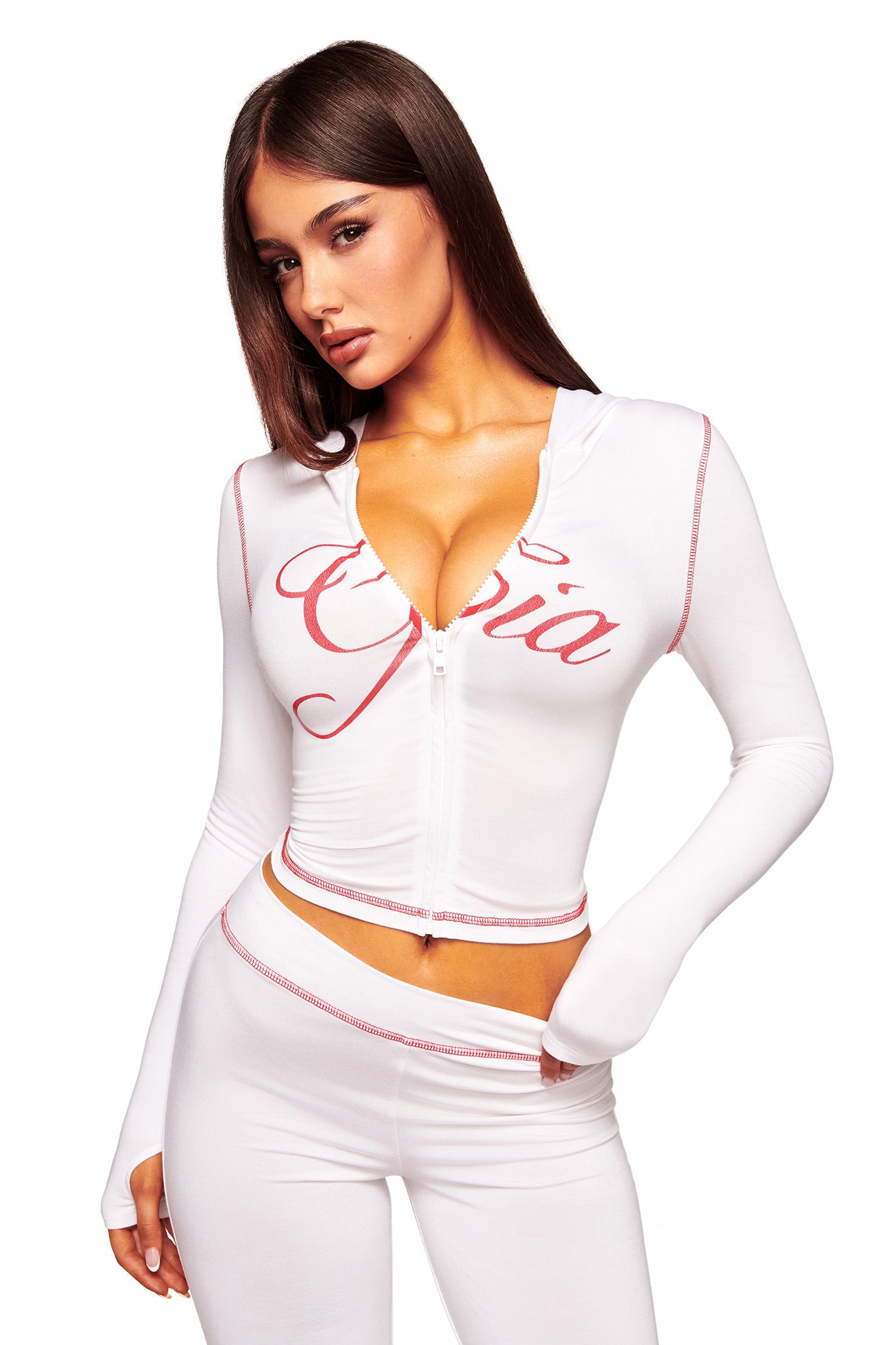 A woman with long brown hair models the I.AM.GIA BLARE HOODIE in white, featuring cursive red text, and matching white pants. She poses with one hand on her hip against a plain white background.
