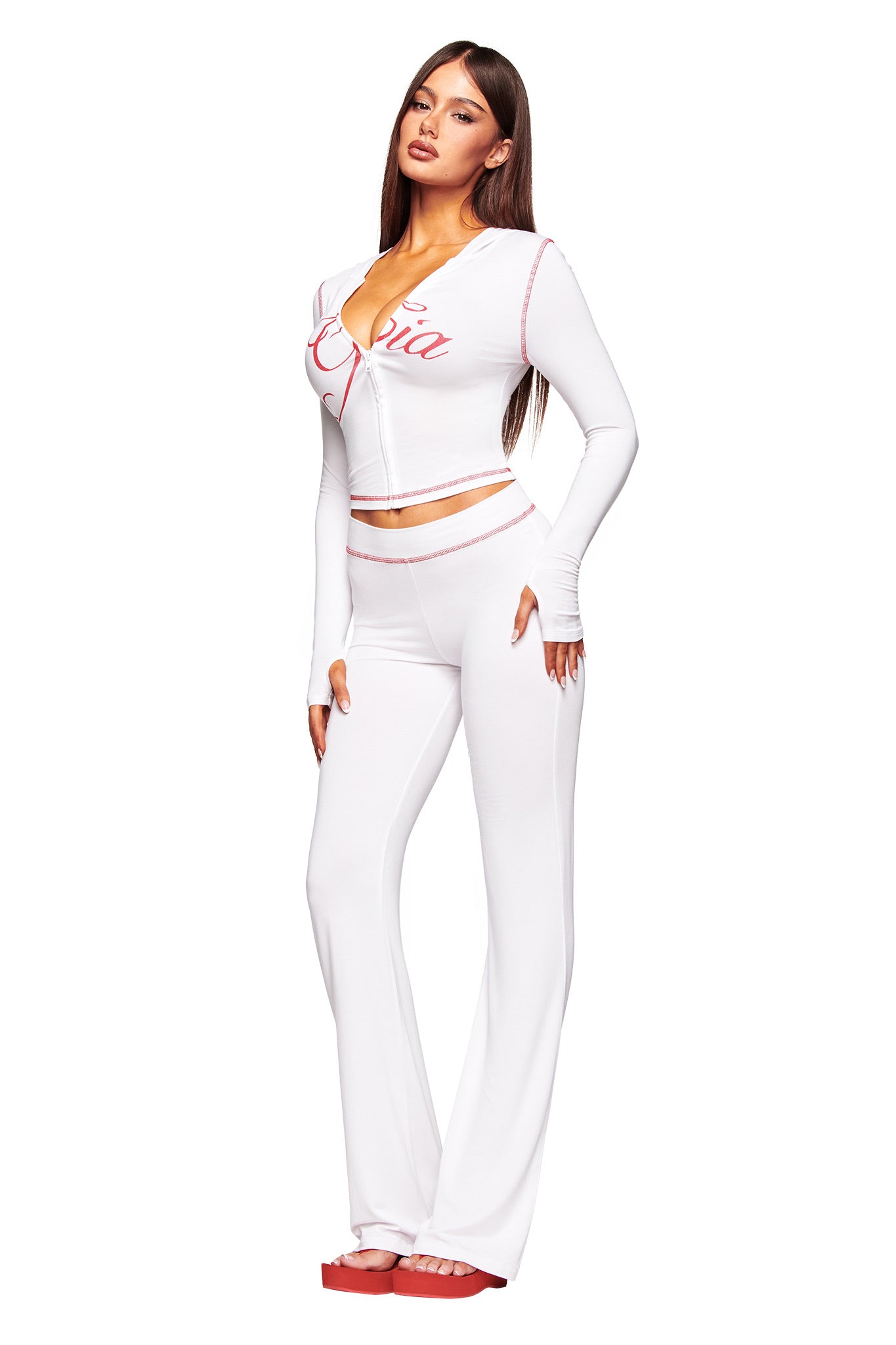 A woman stands against a white background, wearing a white long-sleeve crop top with red trim and “Coca” text, paired with I.AM.GIA’s BLARE TRACKPANT - WHITE featuring a low-rise waist, and red platform sandals.