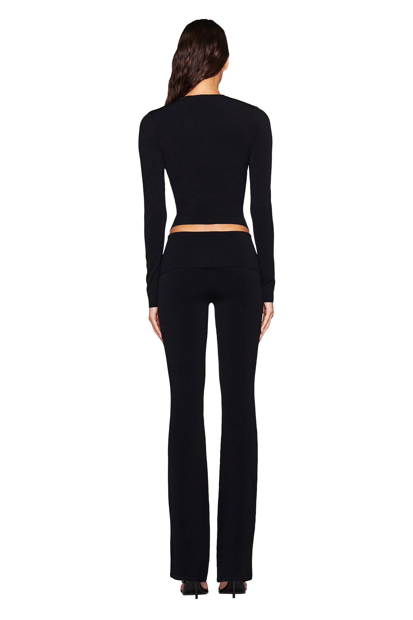 A person with long hair faces away in a black bodycon top and AUDRINA PANT 2.0 - BLACK by I.AM.GIA, featuring a high-waisted fit with a foldover waistband. They stand against a white background.