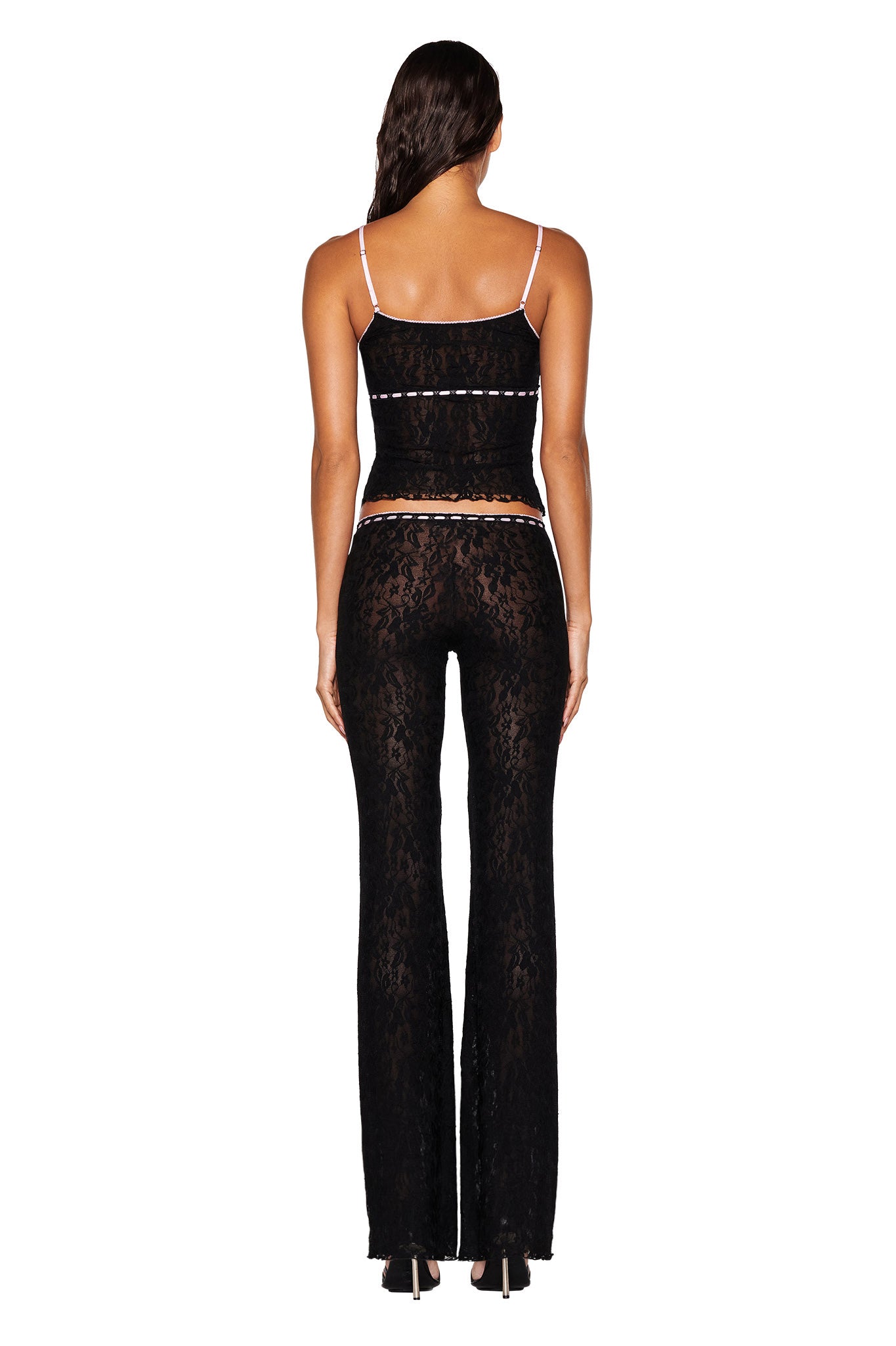 A person with long hair is shown from behind, wearing the LORAH PANT in black by I.AM.GIA, paired with a matching black lace camisole. The outfit features a pull-on construction and showcases sheer floral patterns, white trim, and thin straps that reveal their back and shoulders. They stand with feet slightly apart, with the pants fitted through the hips and flaring at the legs.