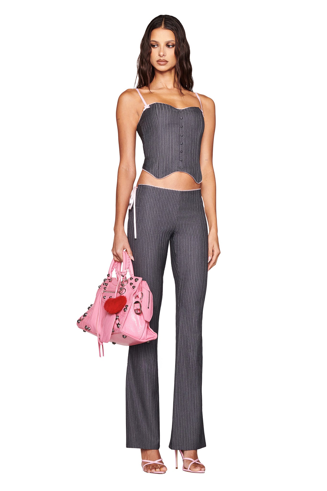 A person is styled in I.AM.GIA's FLORIAN CORSET - GREY, a sleek grey pinstripe ensemble with matching pants. They accessorize with a pink handbag adorned with heart patches and wear strappy heels. Their long, wavy hair cascades elegantly over their shoulders, enhancing the chic vibe.