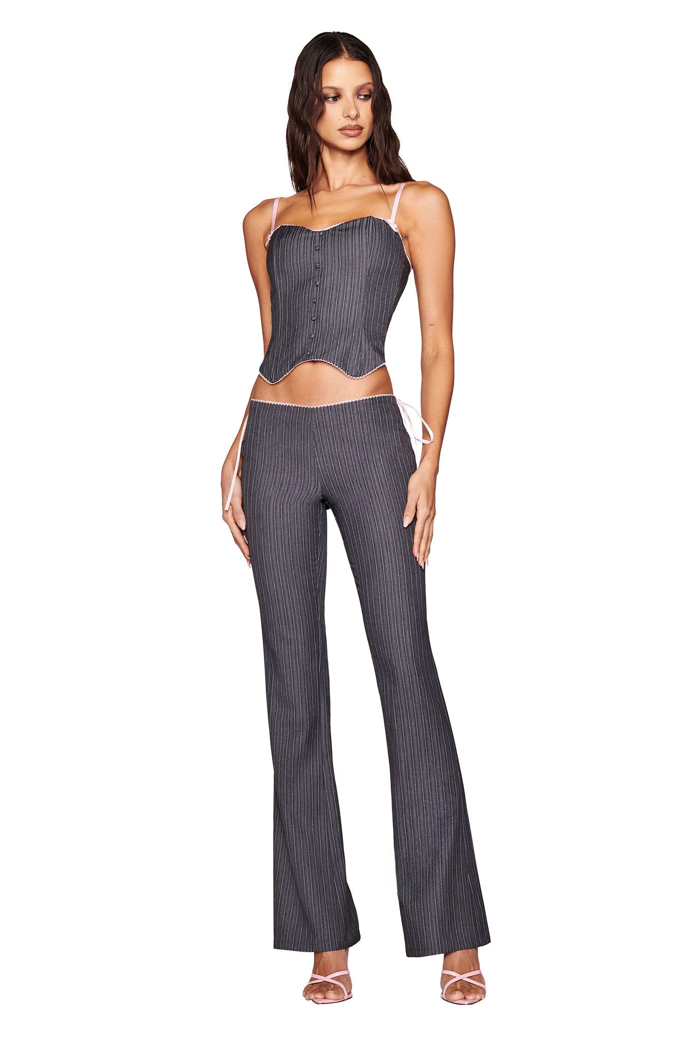 A woman in the I.AM.GIA FLORIAN CORSET - GREY: GREY PINSTRIPE, featuring a tailored fit with a sweetheart neckline and matching flared pants, stands against a white background. She looks forward with long hair cascading down and complements her outfit with strappy heels, arms by her sides.