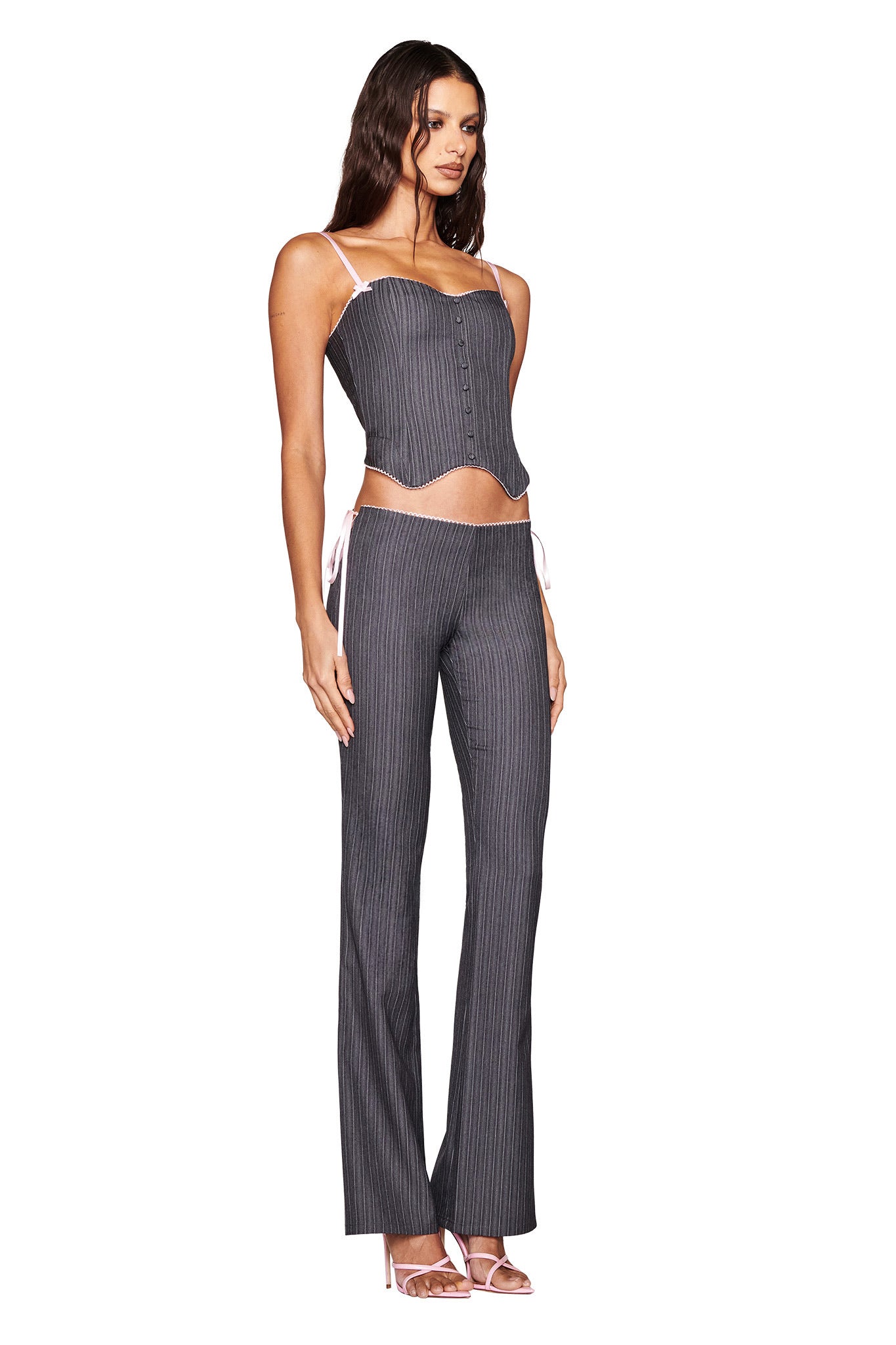 A woman confidently dons the FLORIAN CORSET - GREY from I.AM.GIA, featuring a pinstripe sweetheart neckline. Her long hair flows gracefully, and her look includes flared pants with delicate straps, button details, and white high-heeled sandals.