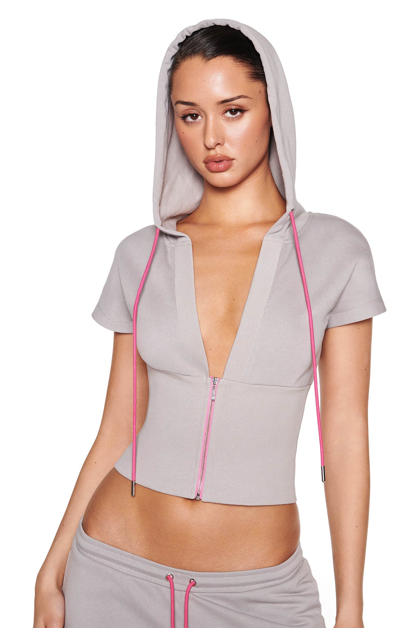 A person stands against a white background wearing an I.AM.GIA ARLO TRACK JACKET in grey, featuring neon pink drawstrings and a deep V-neck. The soft French terry jacket is paired with matching pants incorporating bright pink drawstrings for a cohesive look.