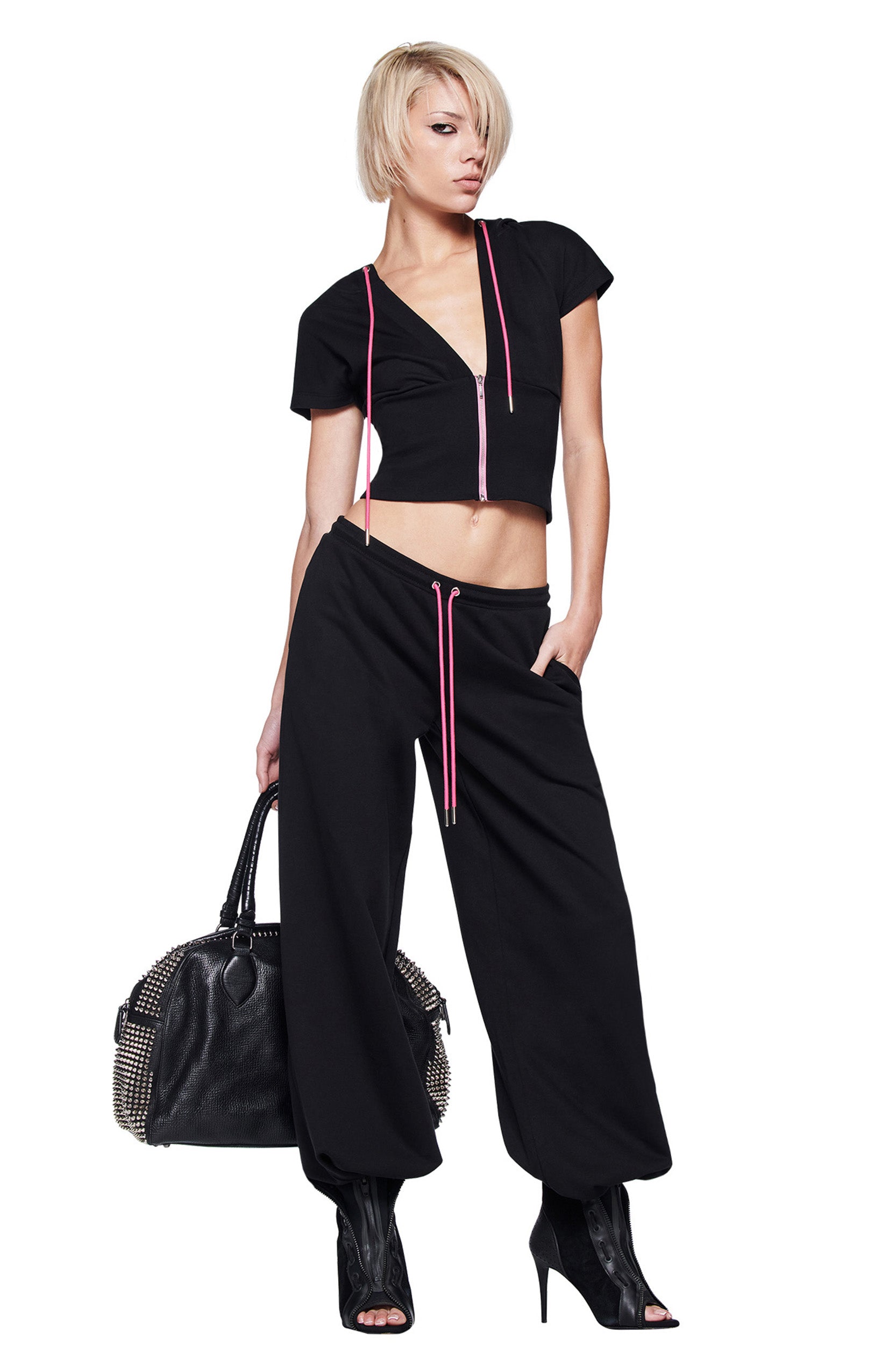 A person with short blond hair wears the I.AM.GIA ARLO TRACK JACKET in black, styled as a cropped top, paired with loose black pants featuring pink drawstrings, a large studded handbag, and black open-toe heeled boots.