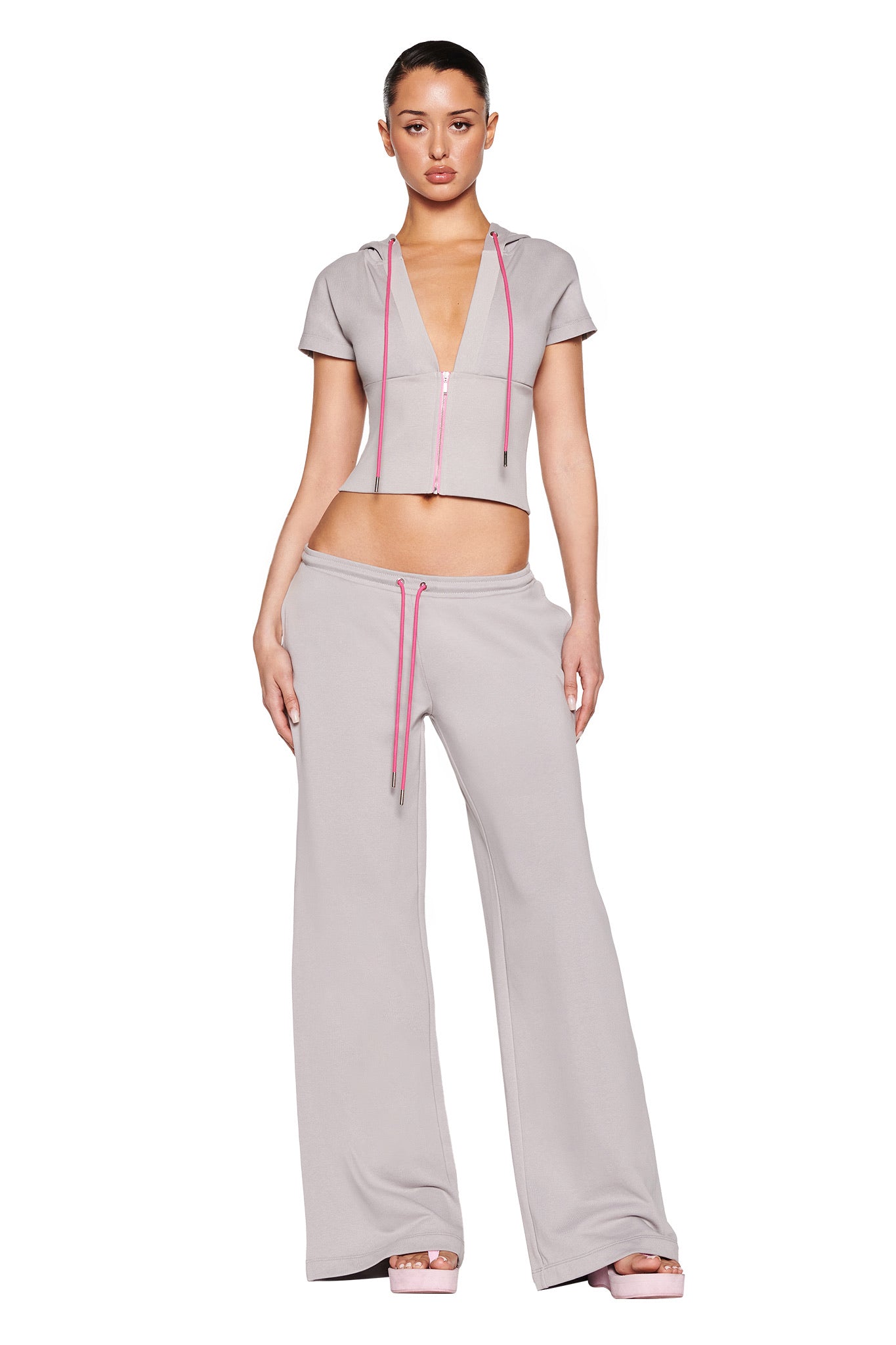 The person is wearing I.AM.GIA's ARLO TRACK PANT in grey with a pink zipper and drawstrings, paired with a light gray, short-sleeved hoodie. The outfit features a deep neckline and subtle brand print as they stand against a plain white background.