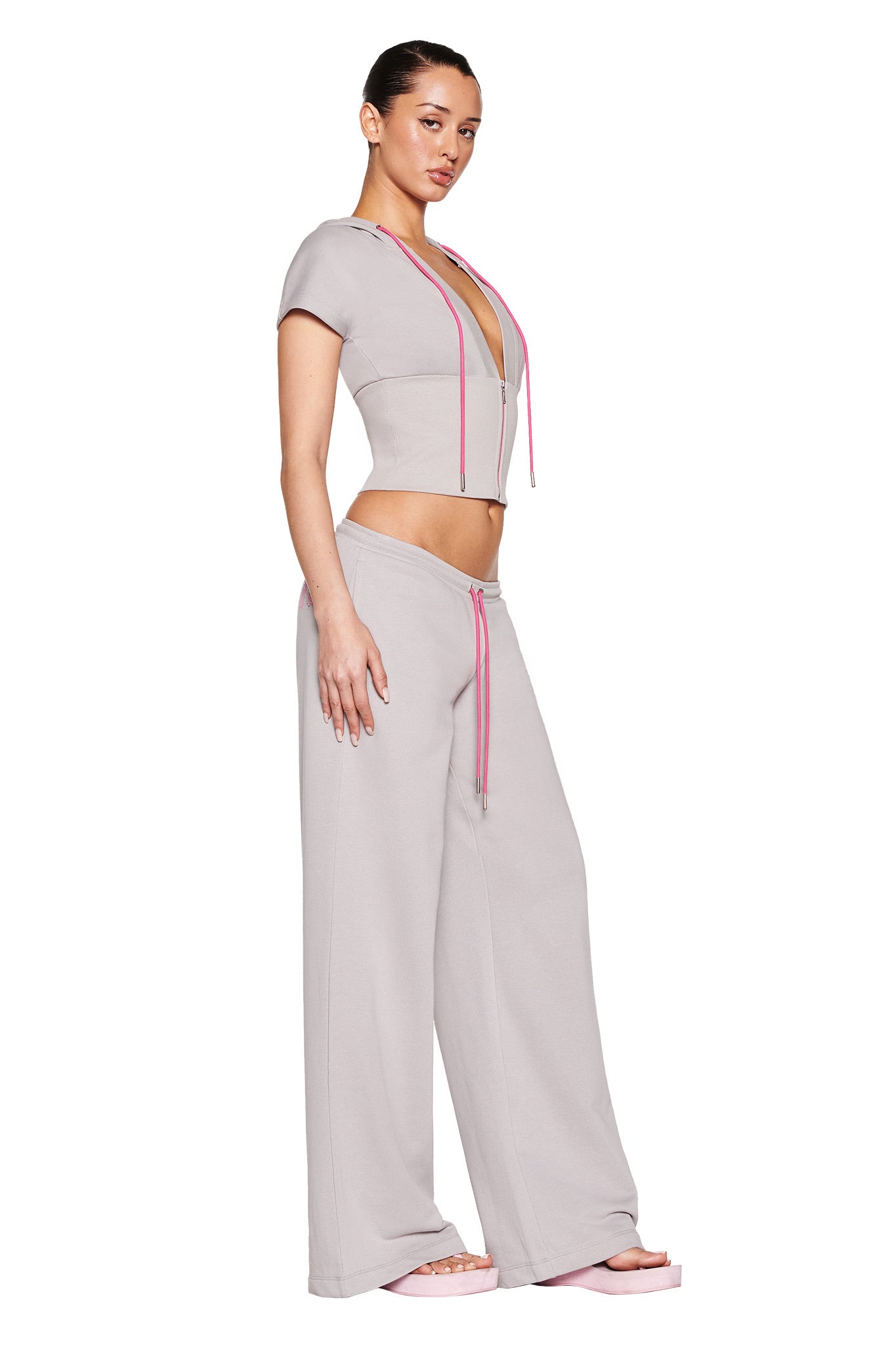 Someone poses in I.AM.GIA's ARLO TRACK PANT in grey with pink accents, paired with a matching short-sleeve top and pink sandals, against a white background.