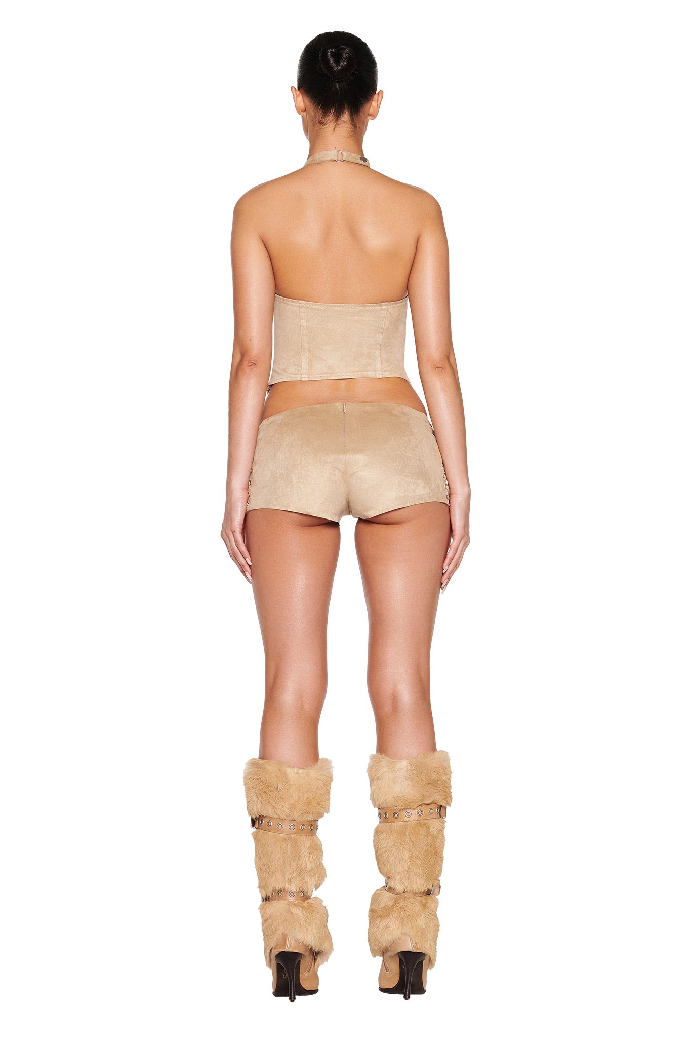 Facing away, a person wears the I.AM.GIA ZEPHYR TOP in beige/tan with matching bodycon shorts and fur-covered leg warmers paired with high heels. Their hair is elegantly styled in a bun against a white background.