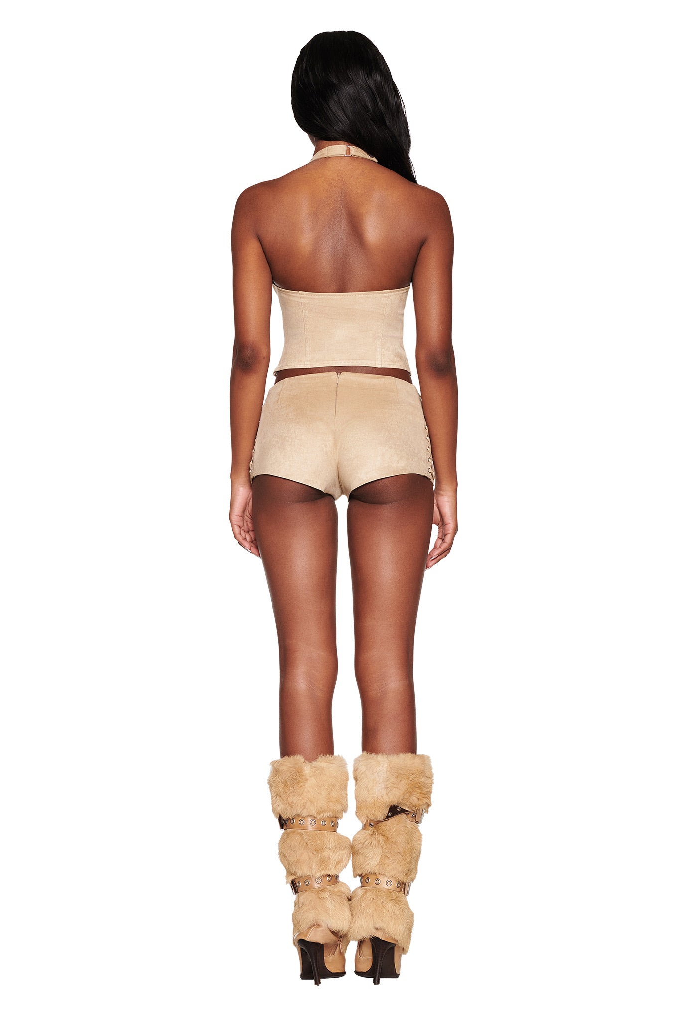 A person with long dark hair is seen from behind in a plain white setting, wearing I.AM.GIA's beige RAMONA SHORT - BEIGE : TAN strapless top, low-rise hot shorts, and tan fur-lined heeled boots.