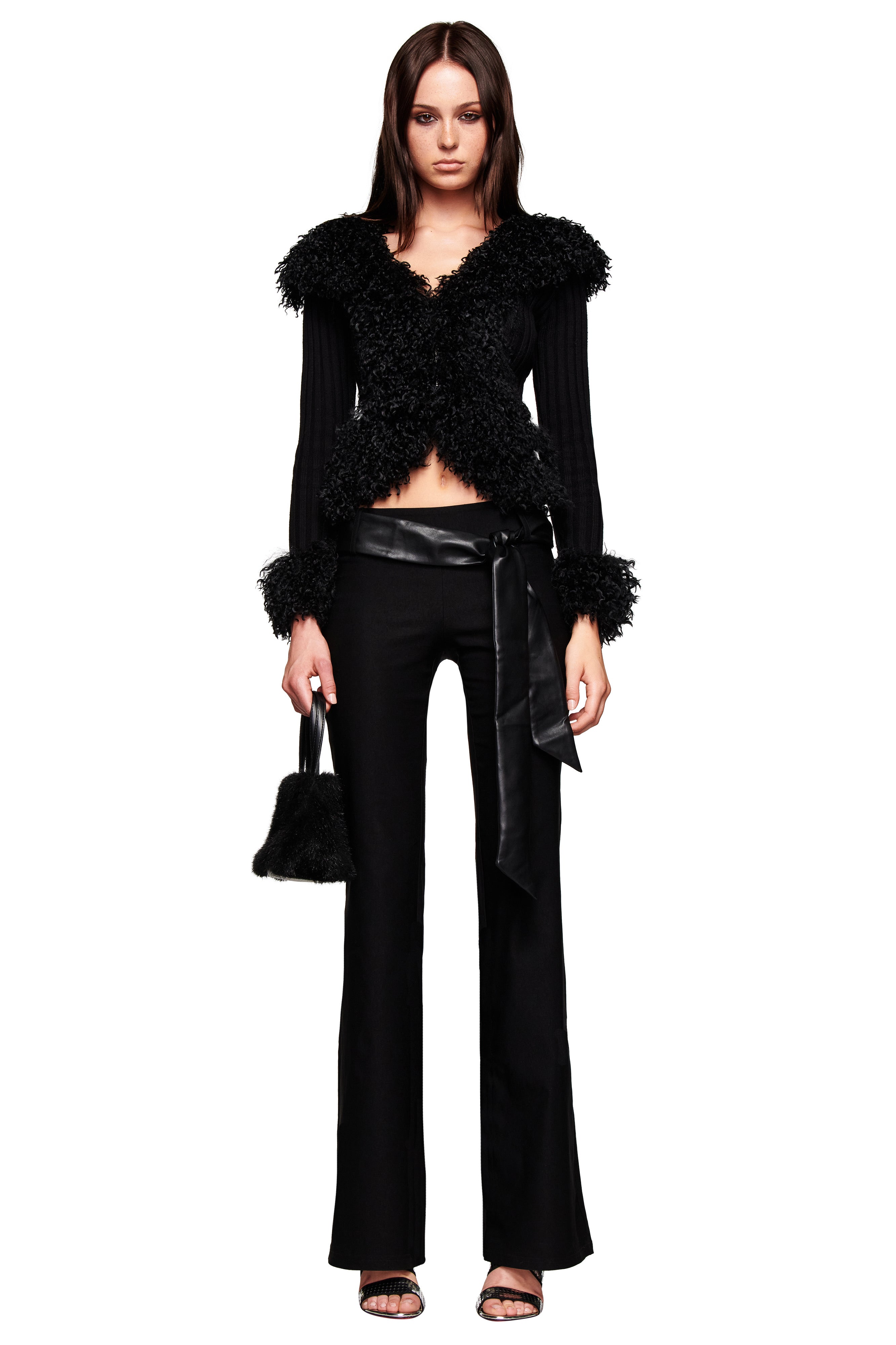 A woman with straight brown hair and a neutral expression wears the I.AM.GIA FELIZ PANT in black, a textured black jacket with fur-like details, high-heeled sandals, and holds a small black bag.