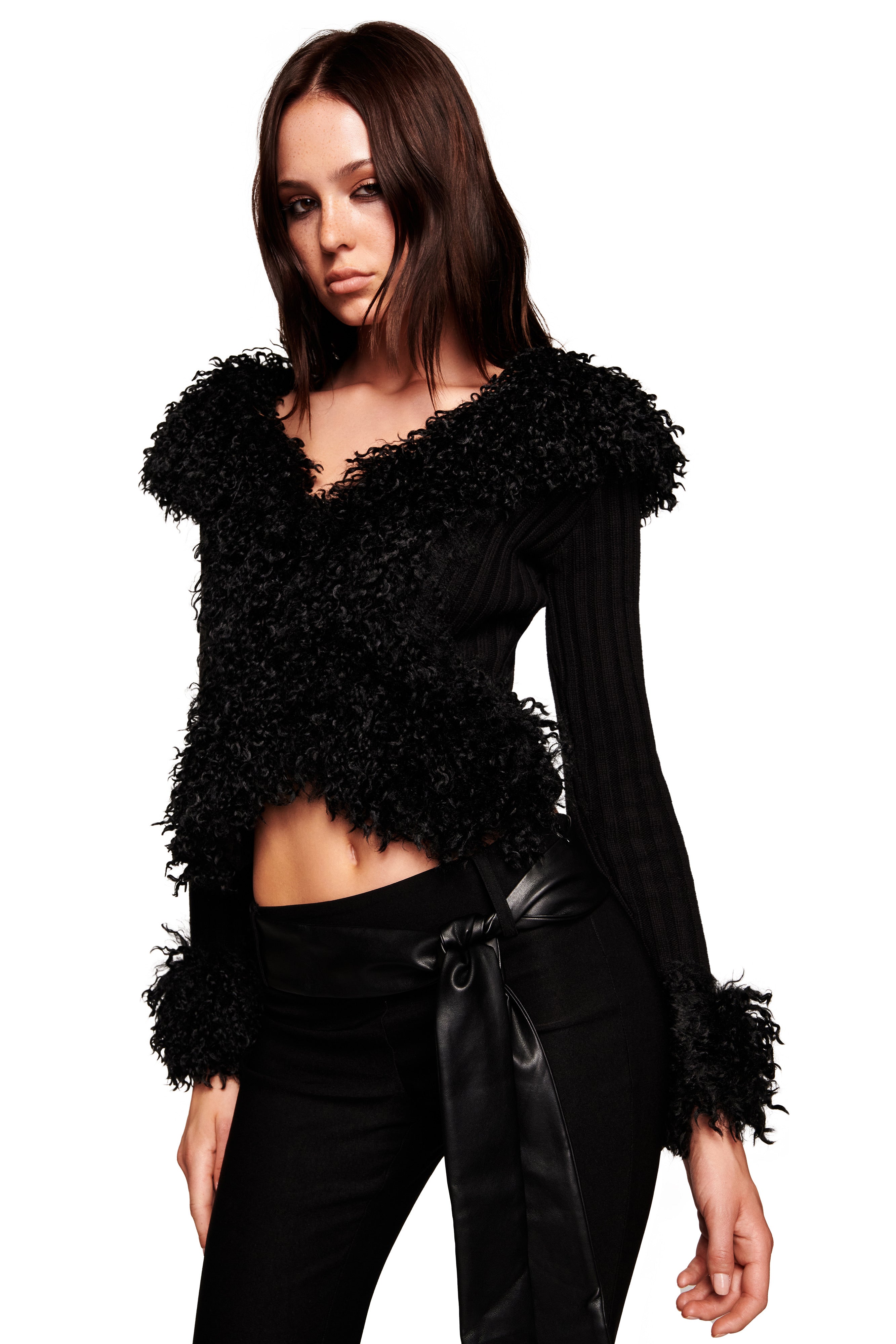 A woman with long brown hair models the I.AM.GIA CRESSIDA JACKET in black, featuring ribbed knit and fluffy details, paired with black pants with a wide satin belt against a white background.