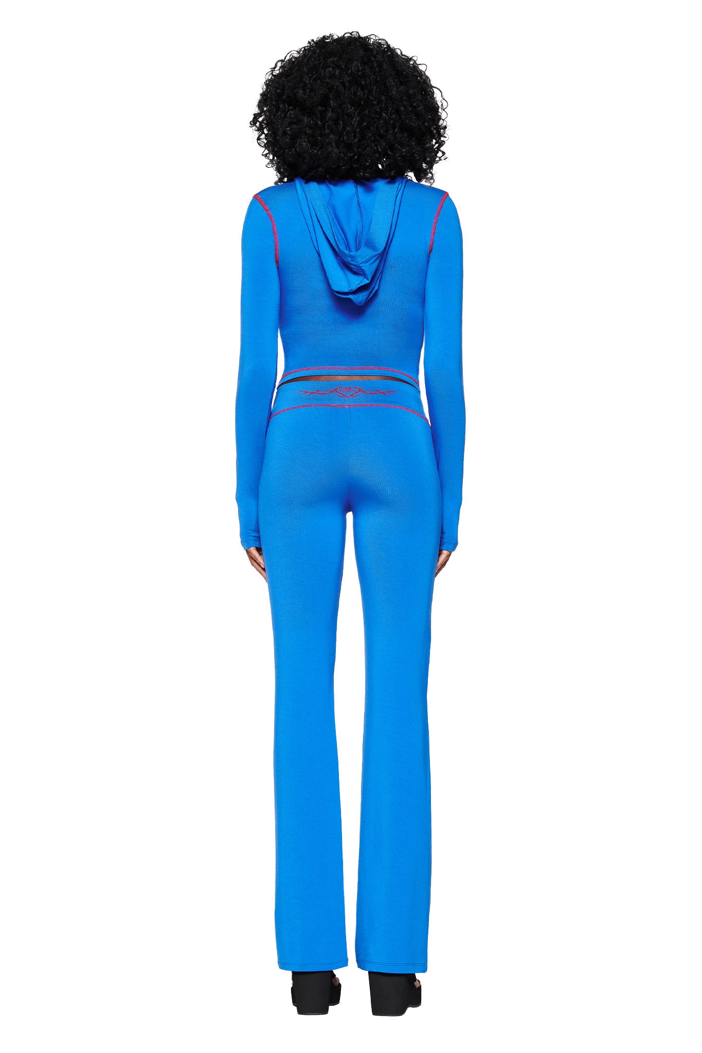 A person with curly hair poses in the I.AM.GIA BLARE HOODIE - BLUE, a royal blue jumpsuit with red accents and mesh waist detail, against a white background. Wearing black shoes, they stand with legs slightly apart.