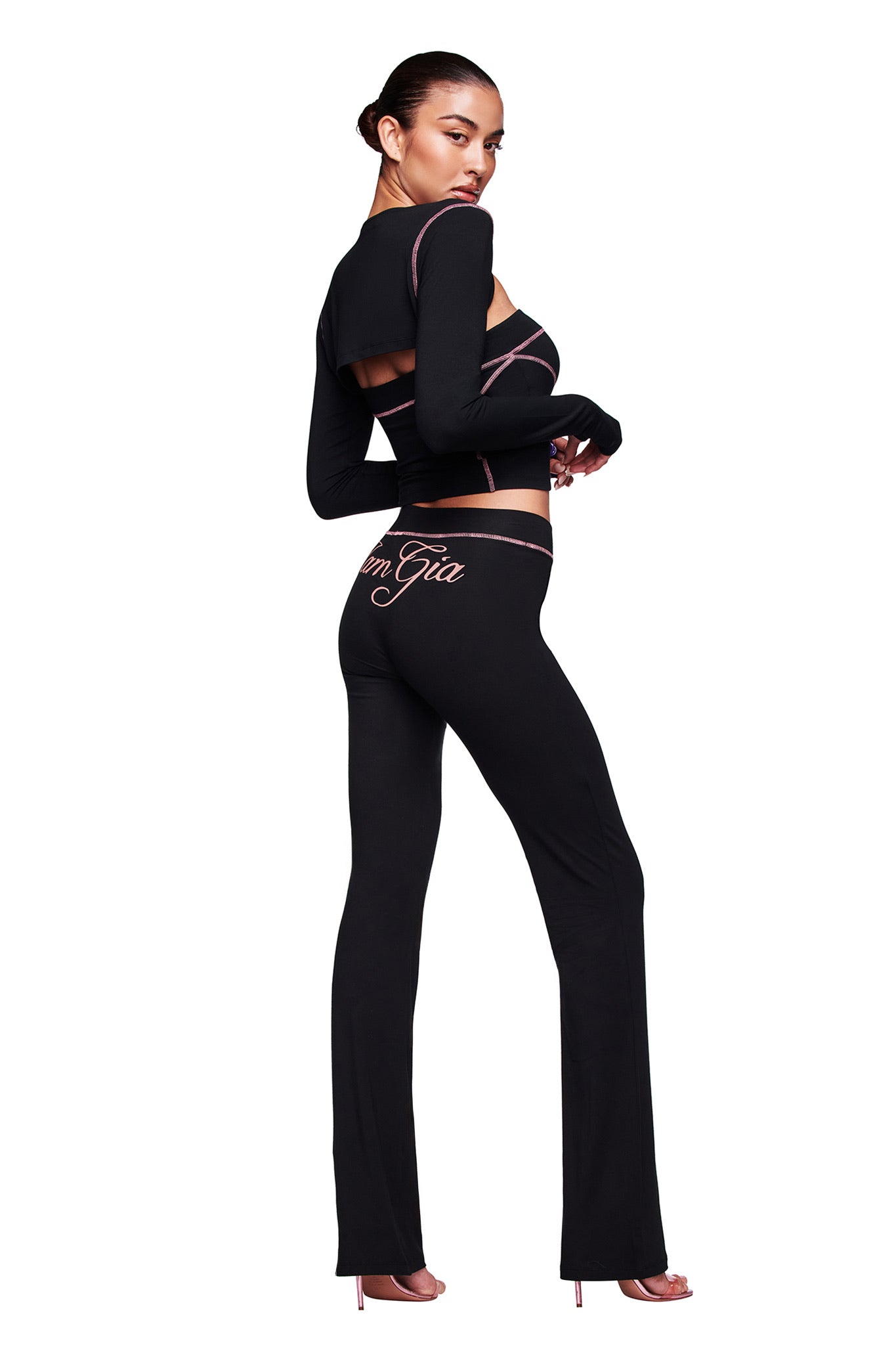 A woman poses confidently in the I.AM.GIA BLARE BOOB TUBE - BLACK, a fitted black two-piece with pink piping and cutout details. The top is strapless, and “I am Gia” appears on the pants as she stands against a white background in heels.