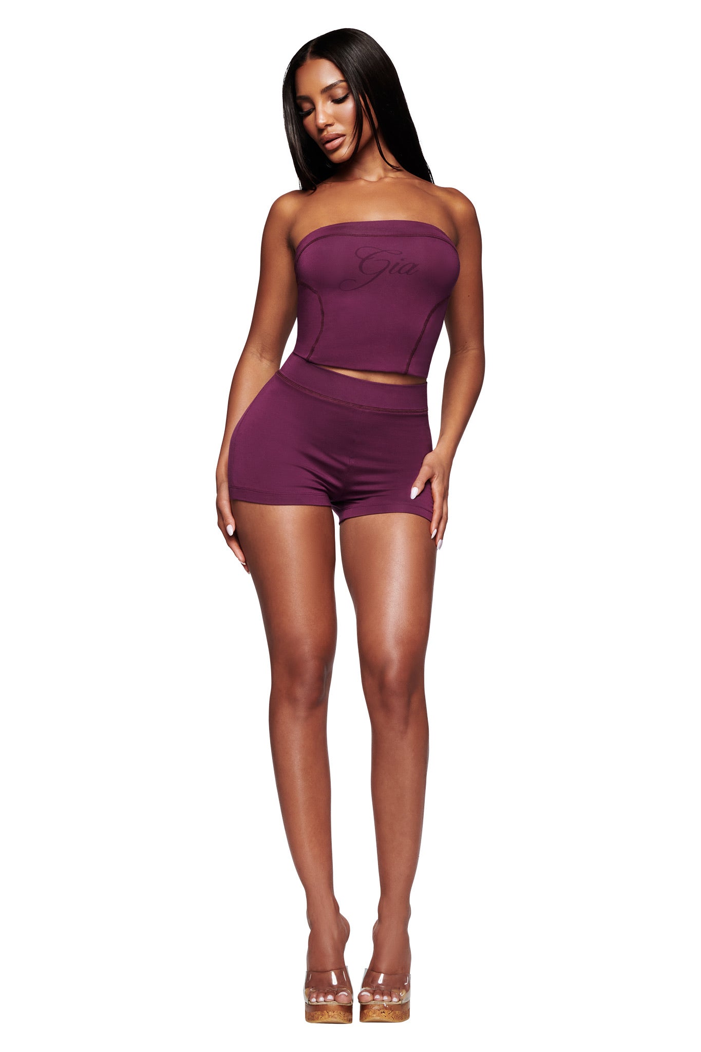 A woman poses against a white background wearing the I.AM.GIA BLARE BOOB TUBE in plum with matching shorts, styled with clear high-heeled shoes.