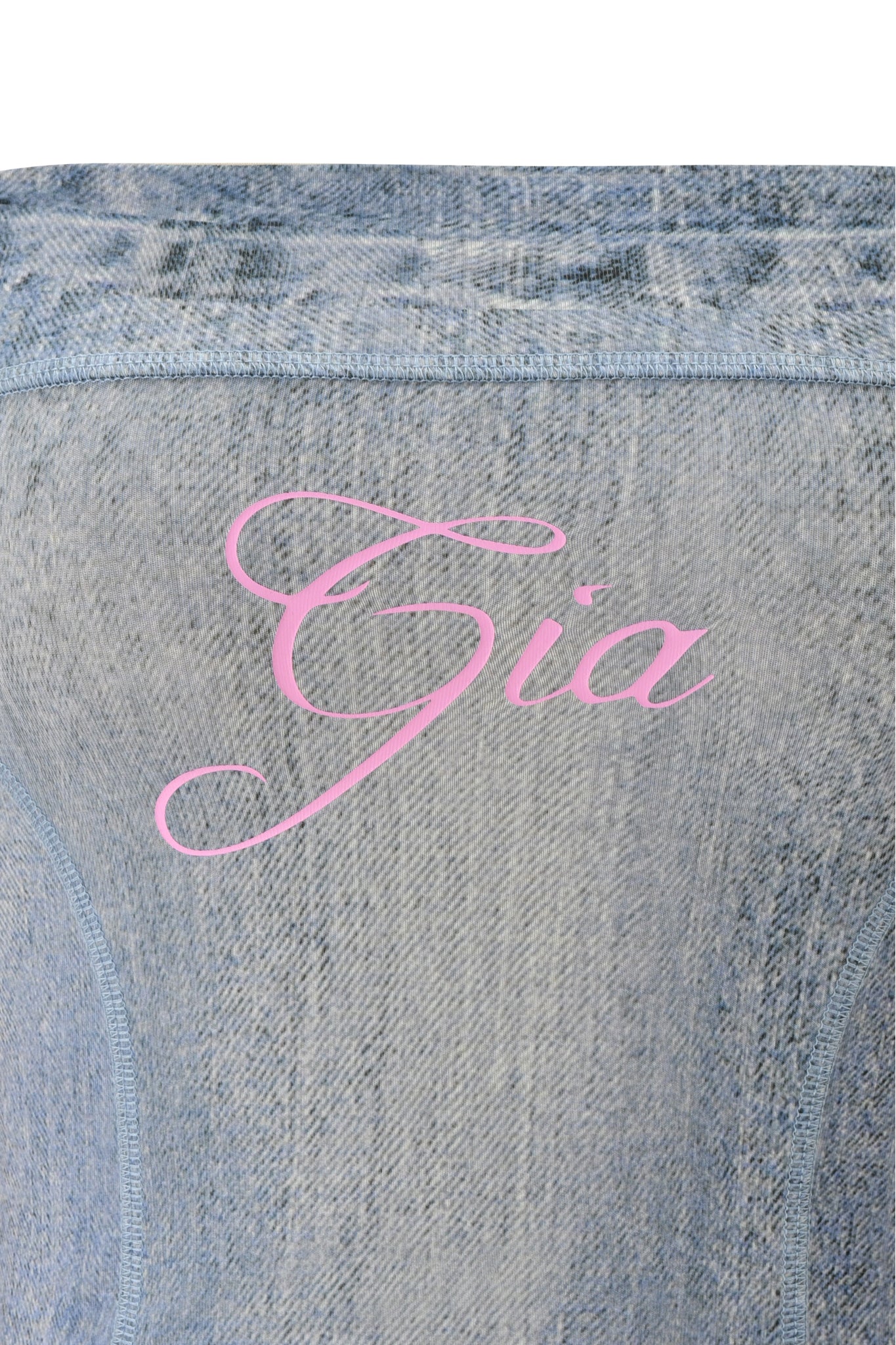 Close-up of light blue denim fabric with "Gia" in elegant pink script, showcasing a subtle trompe l'oeil print inspired by the I.AM.GIA BLARE BOOB TUBE - DENIM TROMPE L'OEIL.