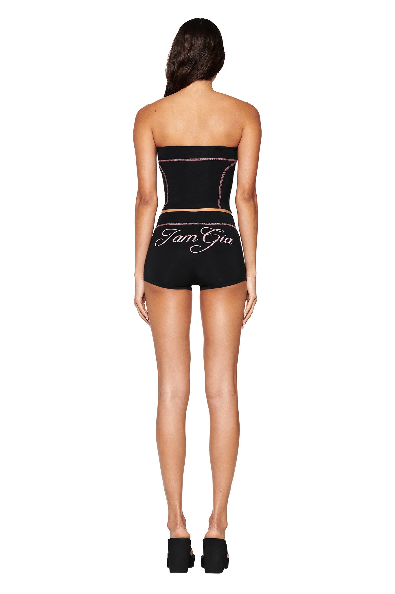 A woman with long hair models the sleek, bodycon fit of I.AM.GIA's 'BLARE SHORT - BLACK,' paired with a black strapless top and black platform shoes against a white background, showcasing "I.AM.GIA" on the back.