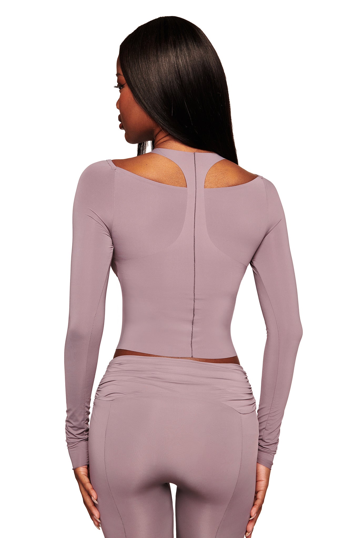A woman stands facing away, wearing I.AM.GIA's NERINA LEGGING in dusty lilac—high-waisted, full-length seamless leggings—paired with a matching fitted, cut-out shoulder lavender crop top for a sporty look.