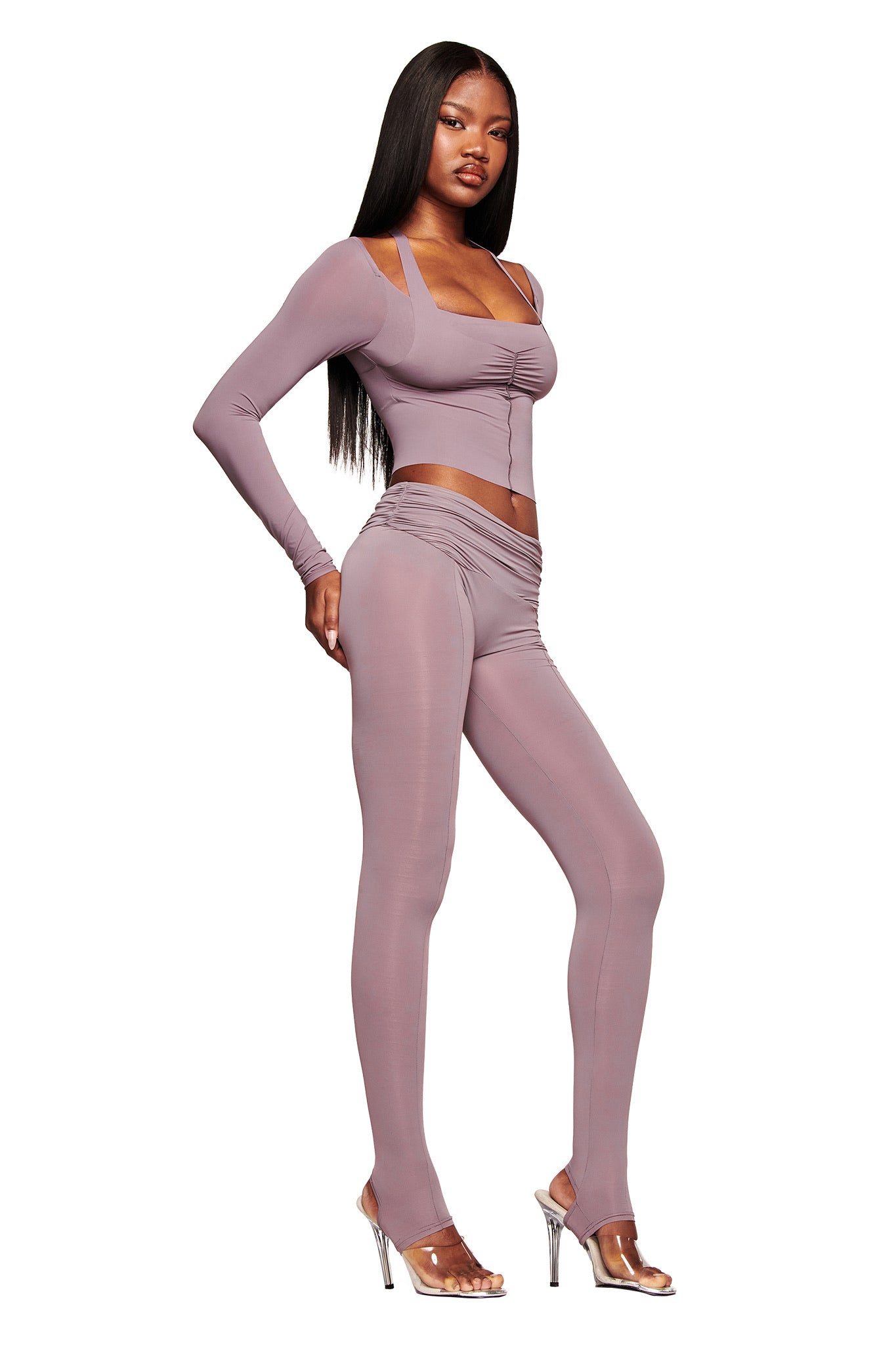 A woman with long straight hair models the I.AM.GIA NERINA LONG SLEEVE TOP in dusty lilac, styled as a bodycon crop top and paired with high-waisted leggings and clear heels against a white background.