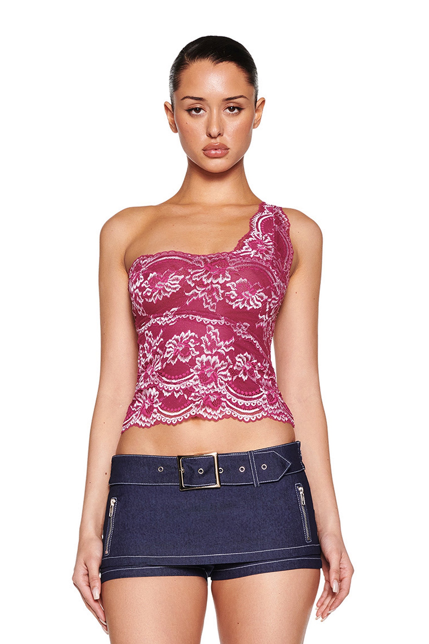 A woman in the I.AM.GIA PHOENIX TOP - PURPLE: PLUM, paired with dark denim shorts featuring zippers, stands against a white background. Her hair is neatly pulled back, accentuating the chic bodycon ensemble.