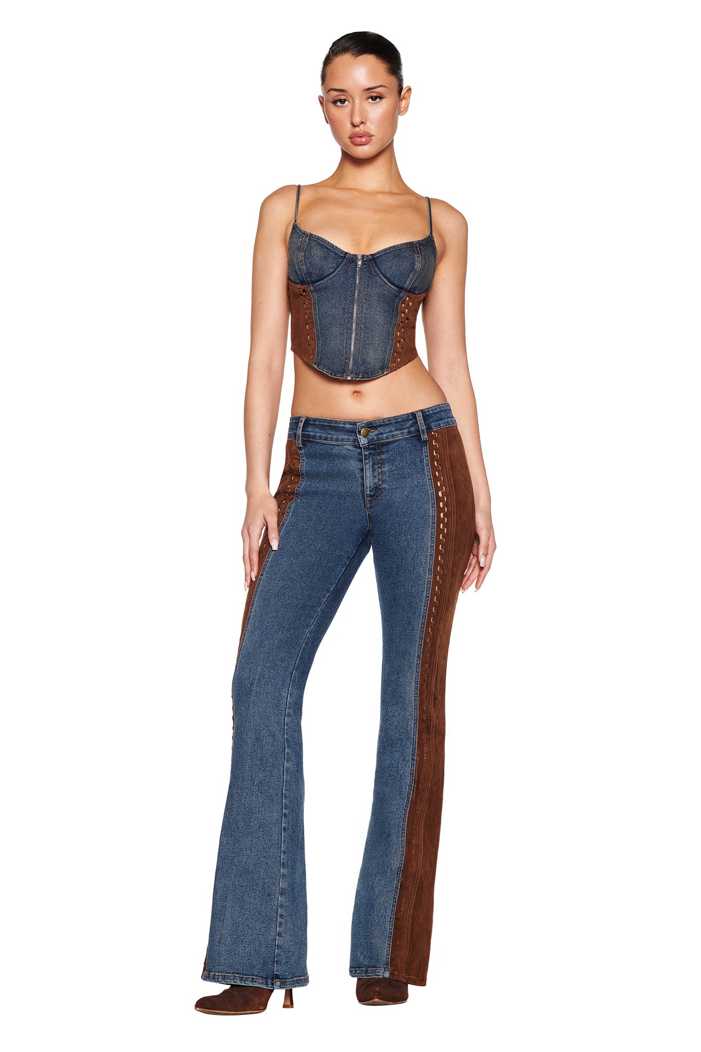 A person poses in a medium wash FORREST PANT by I.AM.GIA, featuring brown suede panels. They wear a denim corset top and brown boots, one hand on their hip and the other relaxed against a plain white background.