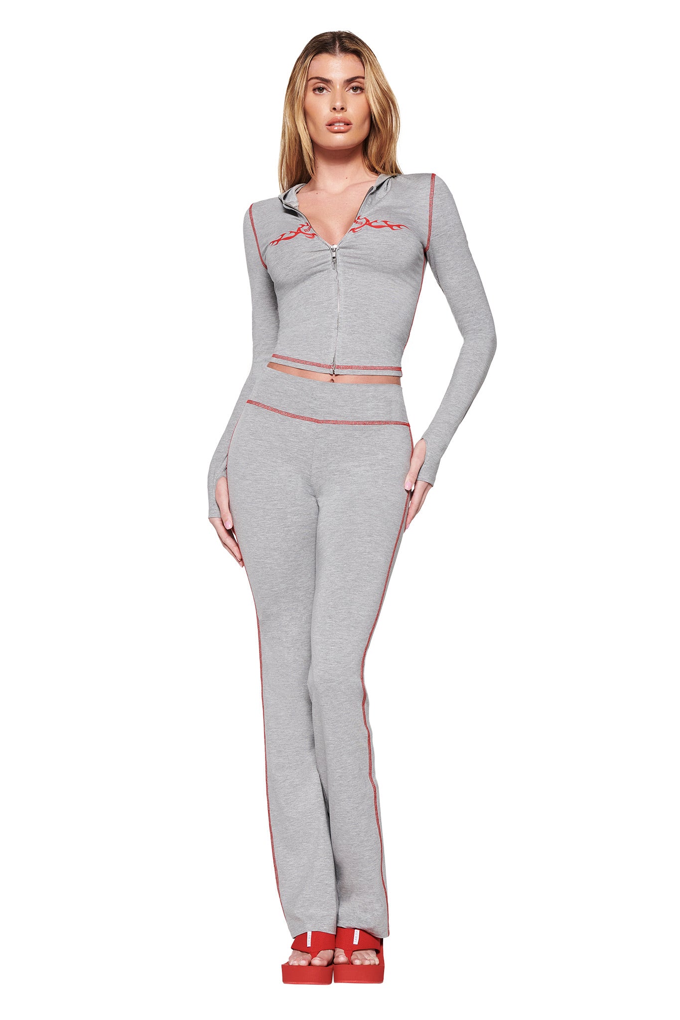 Against a white backdrop, someone models the I.AM.GIA BLARE HOODIE in Dove Grey—a light gray bodycon silhouette with red trim, featuring a zip-up top and wide-leg pants. The ensemble is completed with red platform sandals, embodying the confident style of the GIA Flame logo.