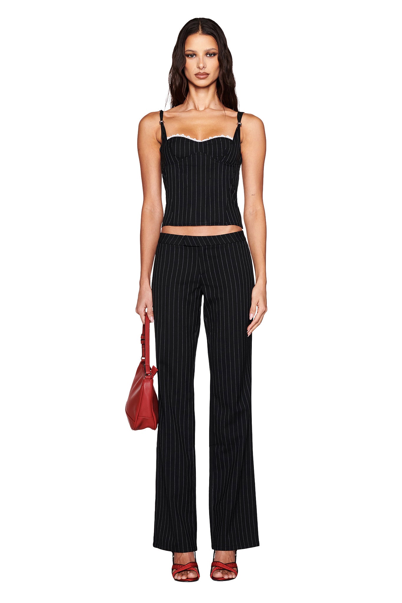 Against a white backdrop, they wear I.AM.GIA's fitted, sleeveless MARRION CORSET in black with lace trim and matching pants. Accessorized with a red handbag and strappy red heels, their long wavy hair completes the look.