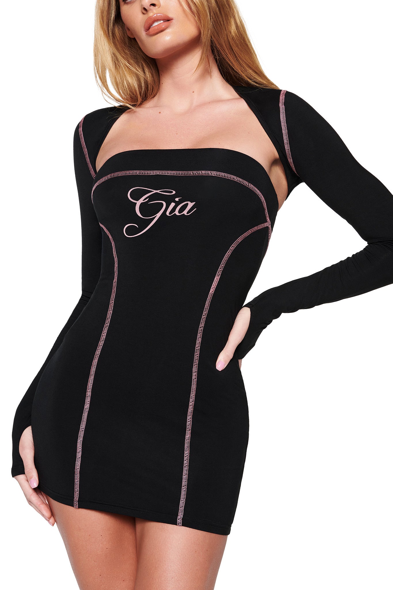 A woman wears a form-fitting black BLARE SHRUG by I.AM.GIA, featuring "Gia" in cursive on the front. Made from ultra-soft stretch bamboo, it showcases long sleeves and pink contrast stitching. She stands confidently with one hand on her hip against a white backdrop.