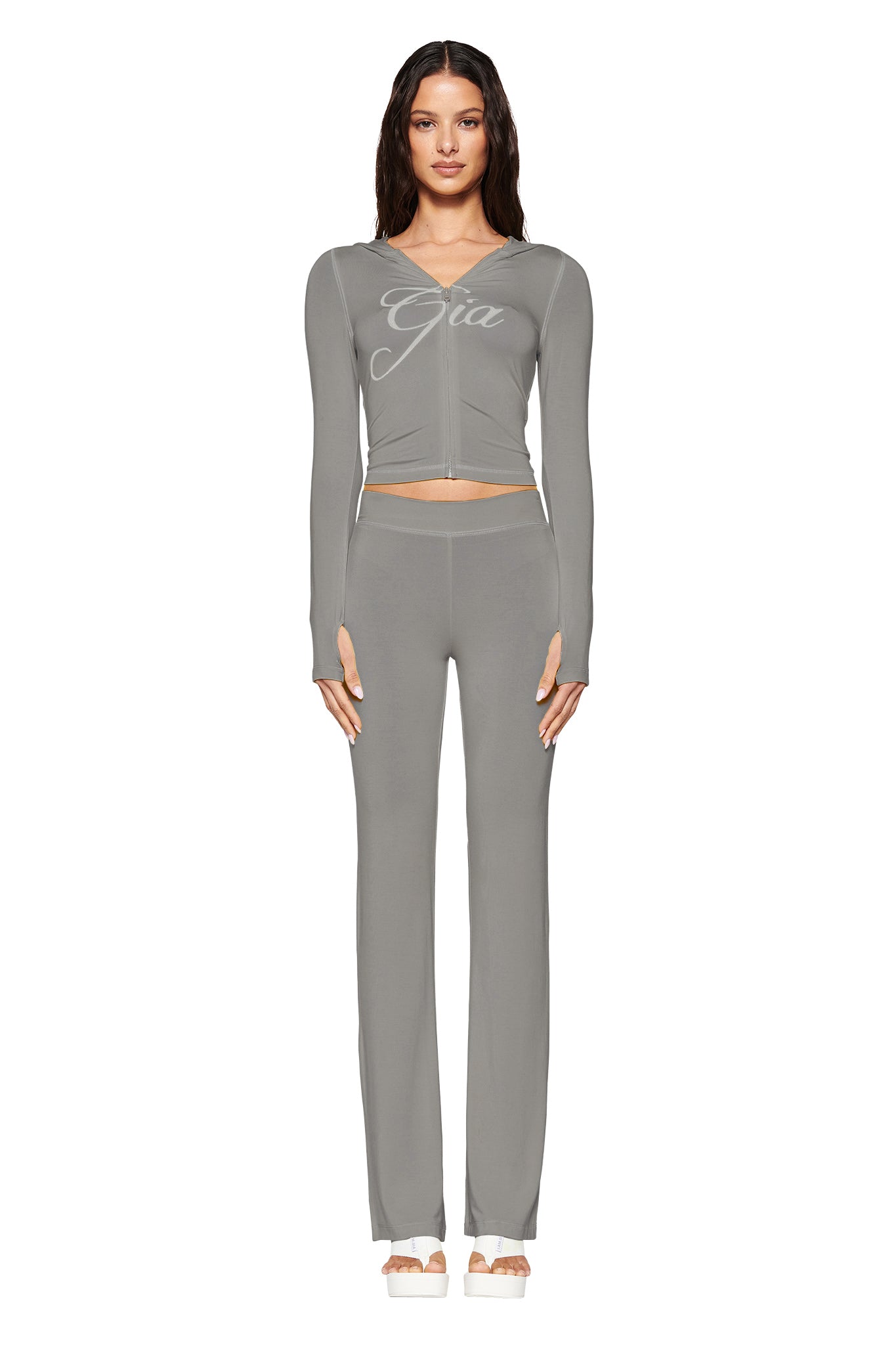 A woman poses in a light gray bodycon outfit featuring a long-sleeve zip-up top and matching pants with "I.AM.GIA" branding. Against a white background, she wears beige shoes and her hair down, embodying effortless elegance.