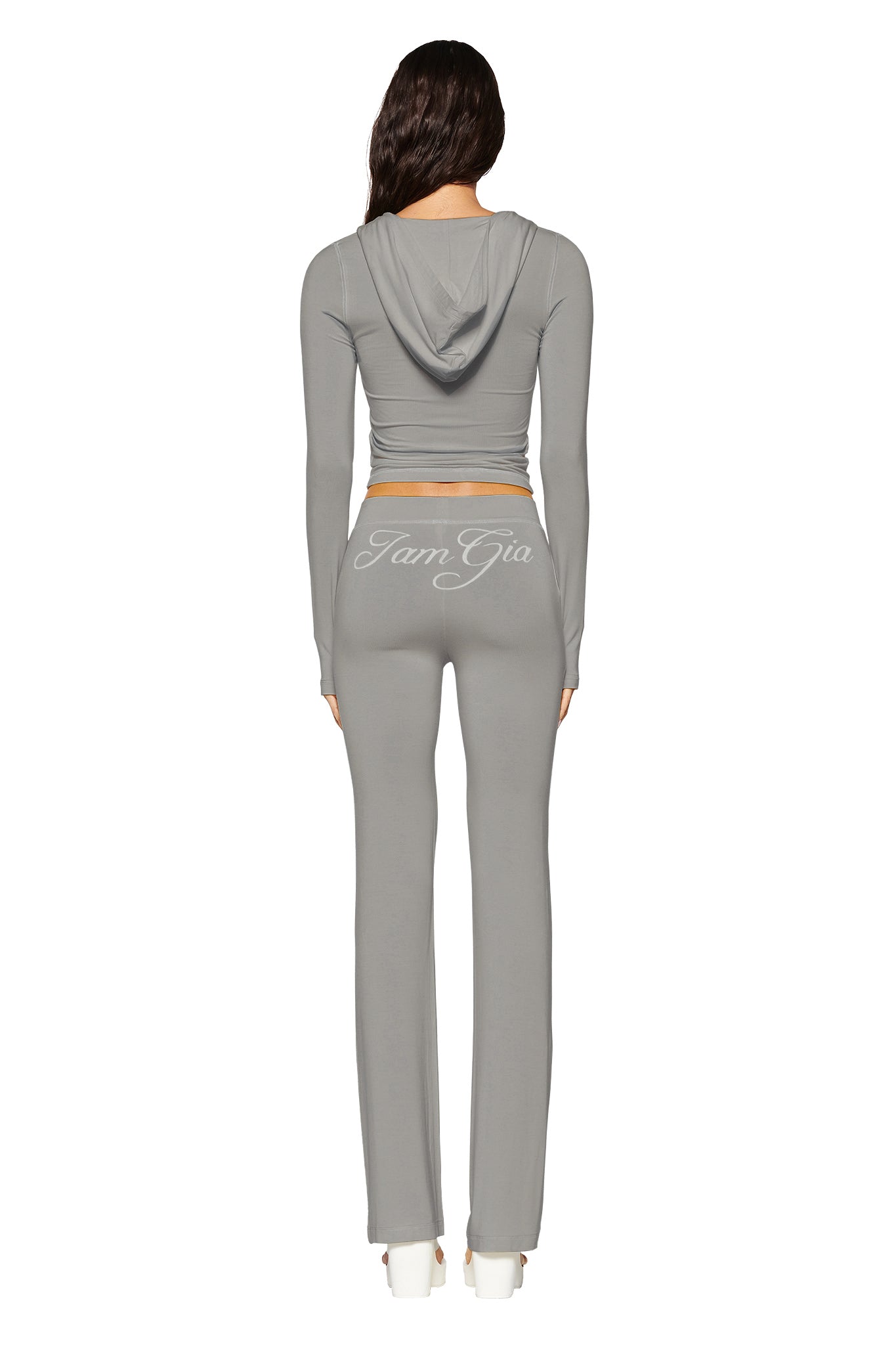 Dressed in a fitted, light gray tracksuit featuring "I.AM.GIA" on the back, a person displays a bodycon silhouette in the Blare Hoodie with flared pants. Set against a white backdrop, they wear white shoes and face away from the camera with effortless style.