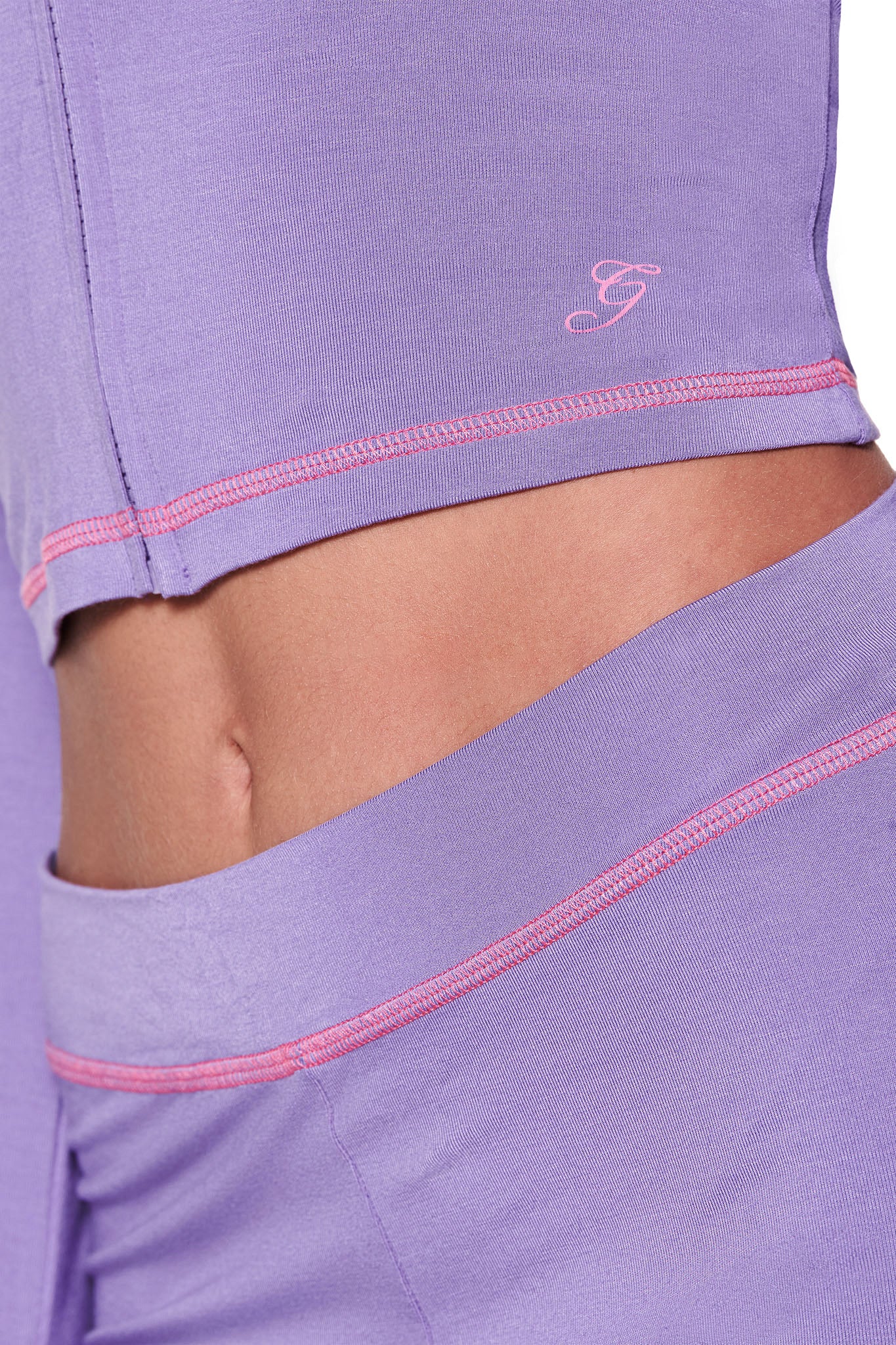 Close-up of a person in I.AM.GIA's BLARE HOODIE - PURPLE, wearing a light purple activewear set with pink stitching. The top is lifted to reveal abs and features a small logo on the waistband. The fabric looks smooth and stretchy.
