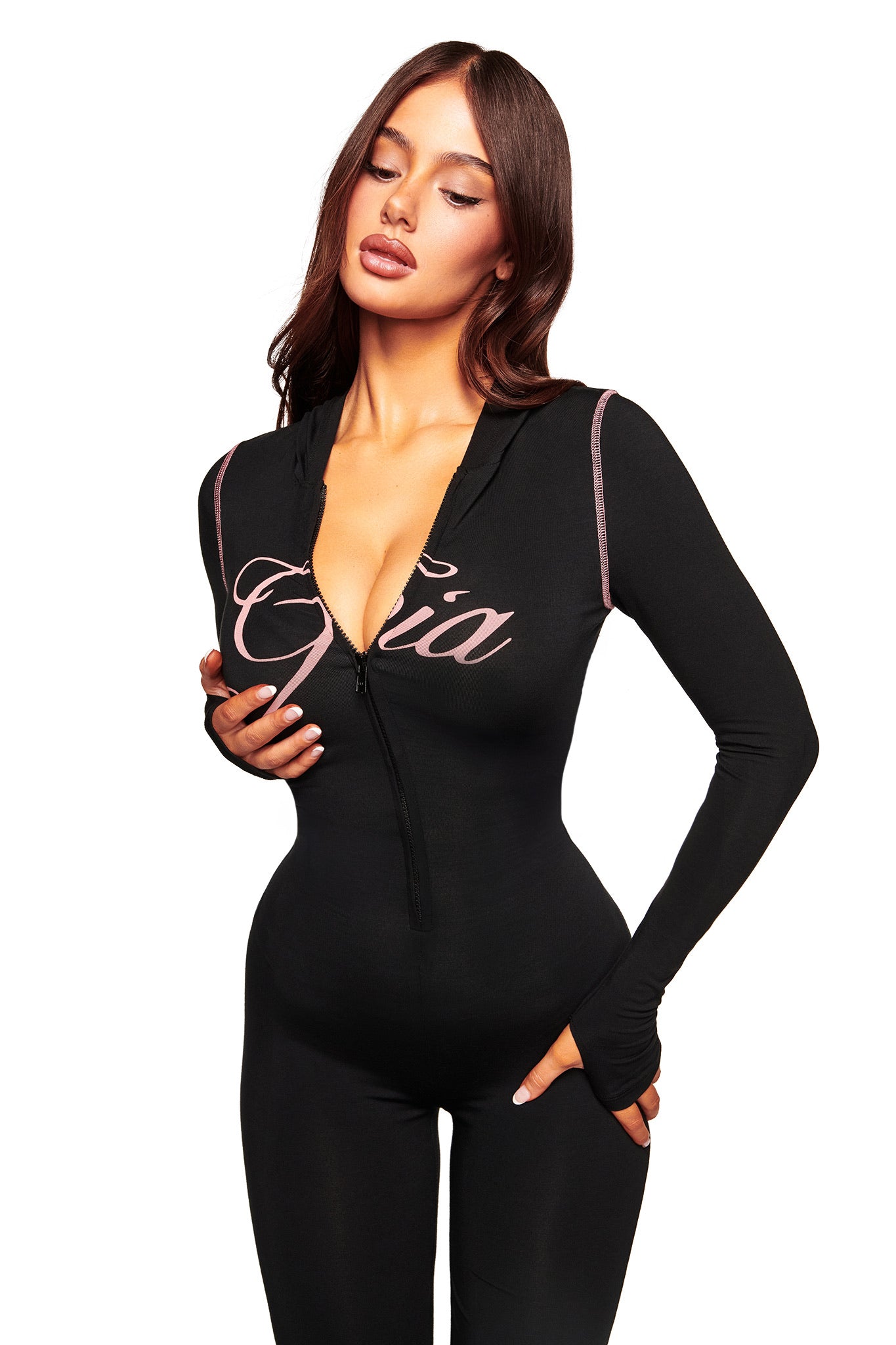 A woman with long brown hair wears the fitted black BLARE JUMPSUIT by I.AM.GIA, featuring a hood, flared legs, and "Gia" script across the chest. She stands against a plain white background, looking down with one hand on her chest.
