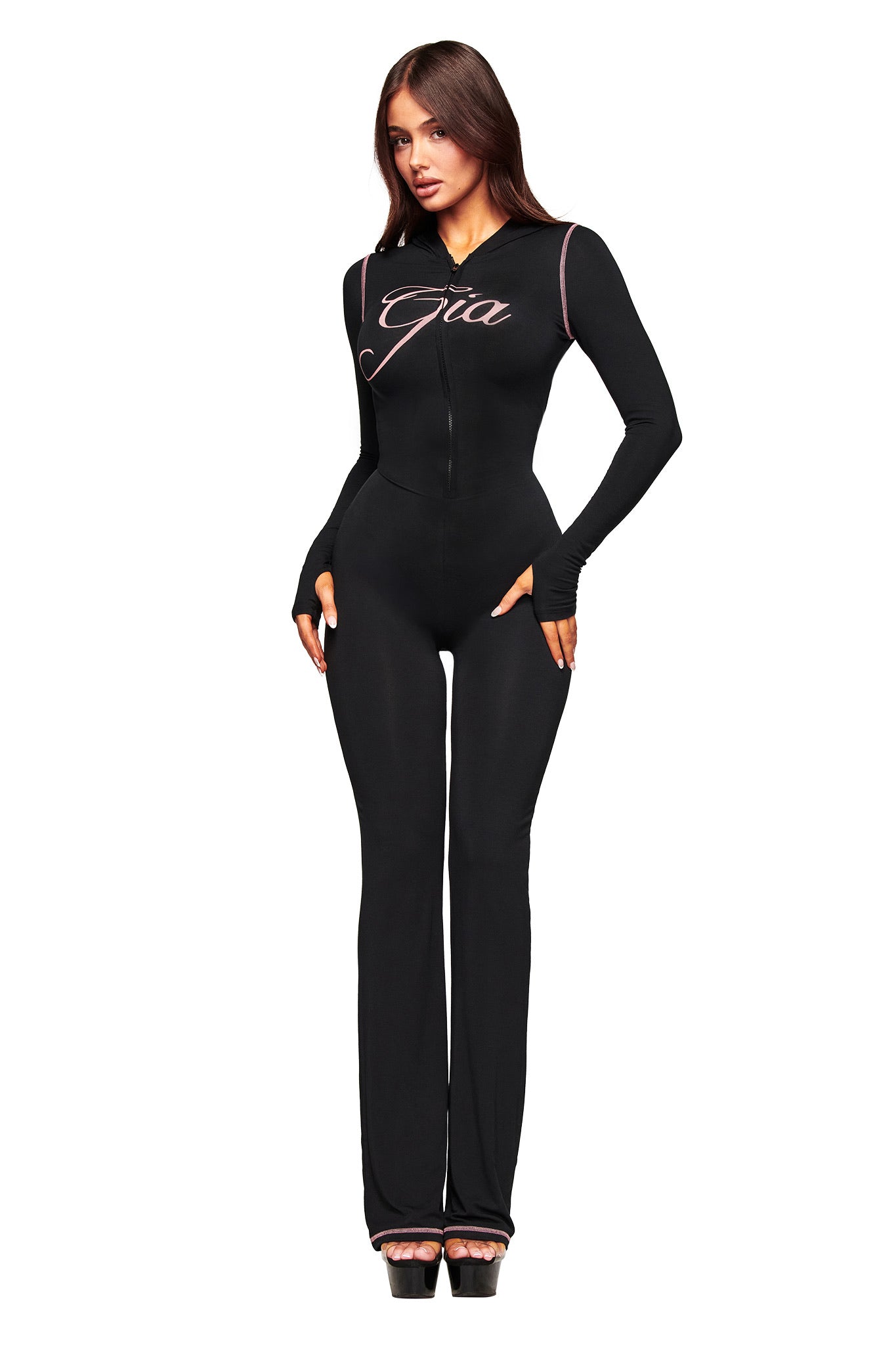 A woman faces forward in the fitted black BLARE JUMPSUIT by I.AM.GIA, featuring "Gia" script on the chest, flared legs, pockets, and paired with black platform heels against a white background.