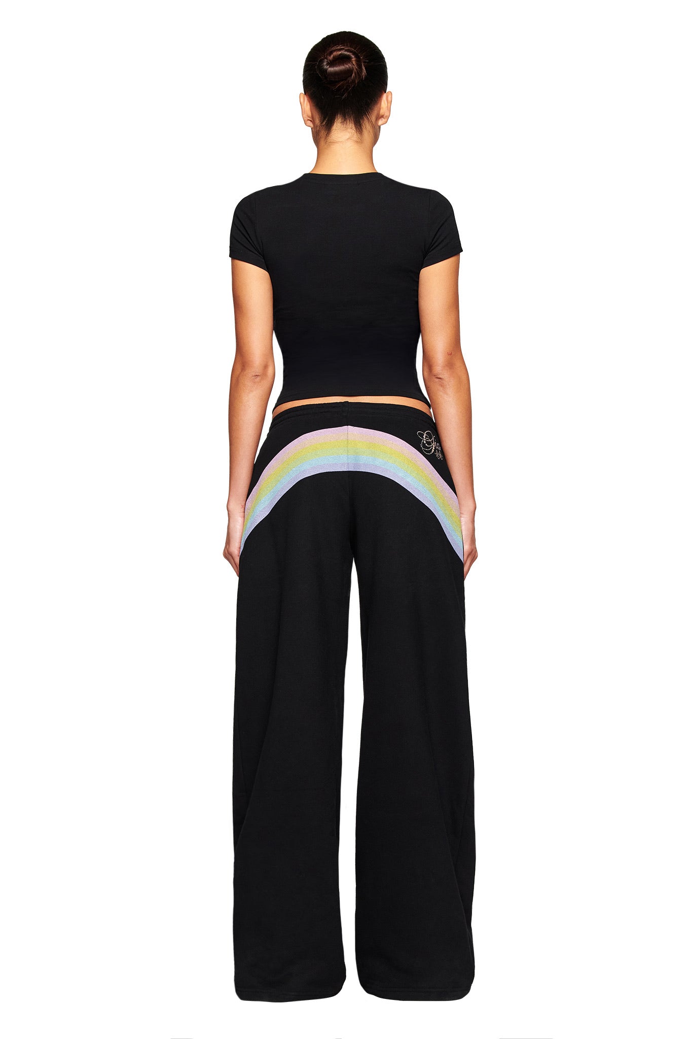 Someone with their hair in a bun is seen from behind wearing the I.AM.GIA FAFI T-SHIRT in black and loose black pants with a bold rainbow design across the back.
