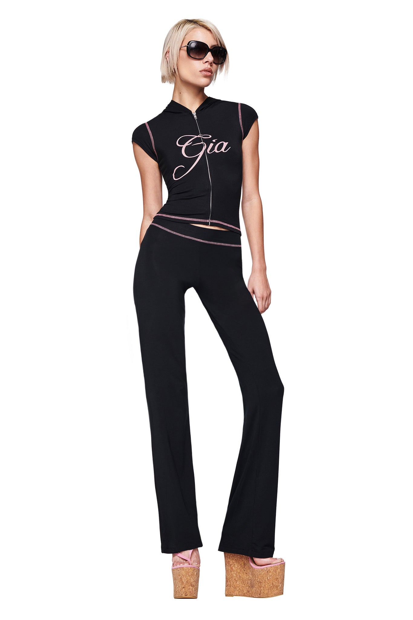 A person with short blonde hair and sunglasses poses confidently against a white background, wearing the I.AM.GIA BLARE CAP SLEEVE HOODIE in black, paired with matching black pants and tan platform sandals.