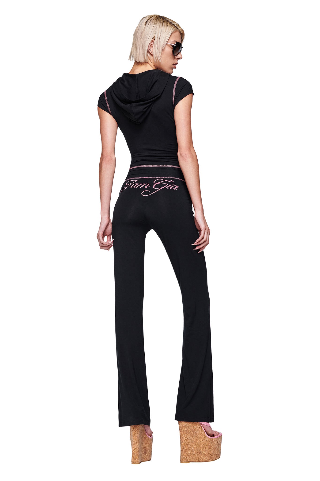A woman with short blonde hair wears sunglasses and a black I.AM.GIA BLARE CAP SLEEVE HOODIE with matching black pants featuring pink writing on the back. Her back is turned, and she pairs the look with high cork platform sandals.