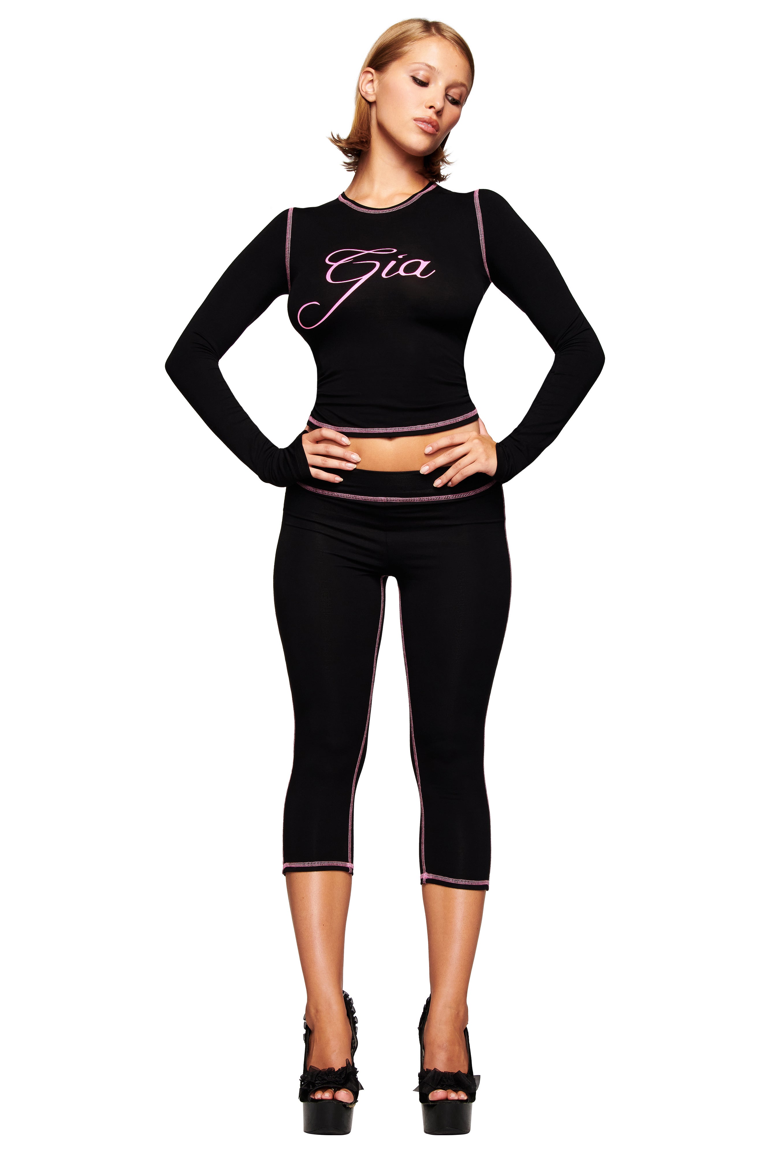 A woman with short blonde hair poses against a white background, wearing the I.AM.GIA BLARE CAPRI - BLACK and a matching black crop top with the brand's logo, styled with high-heeled shoes and her hands on her hips.