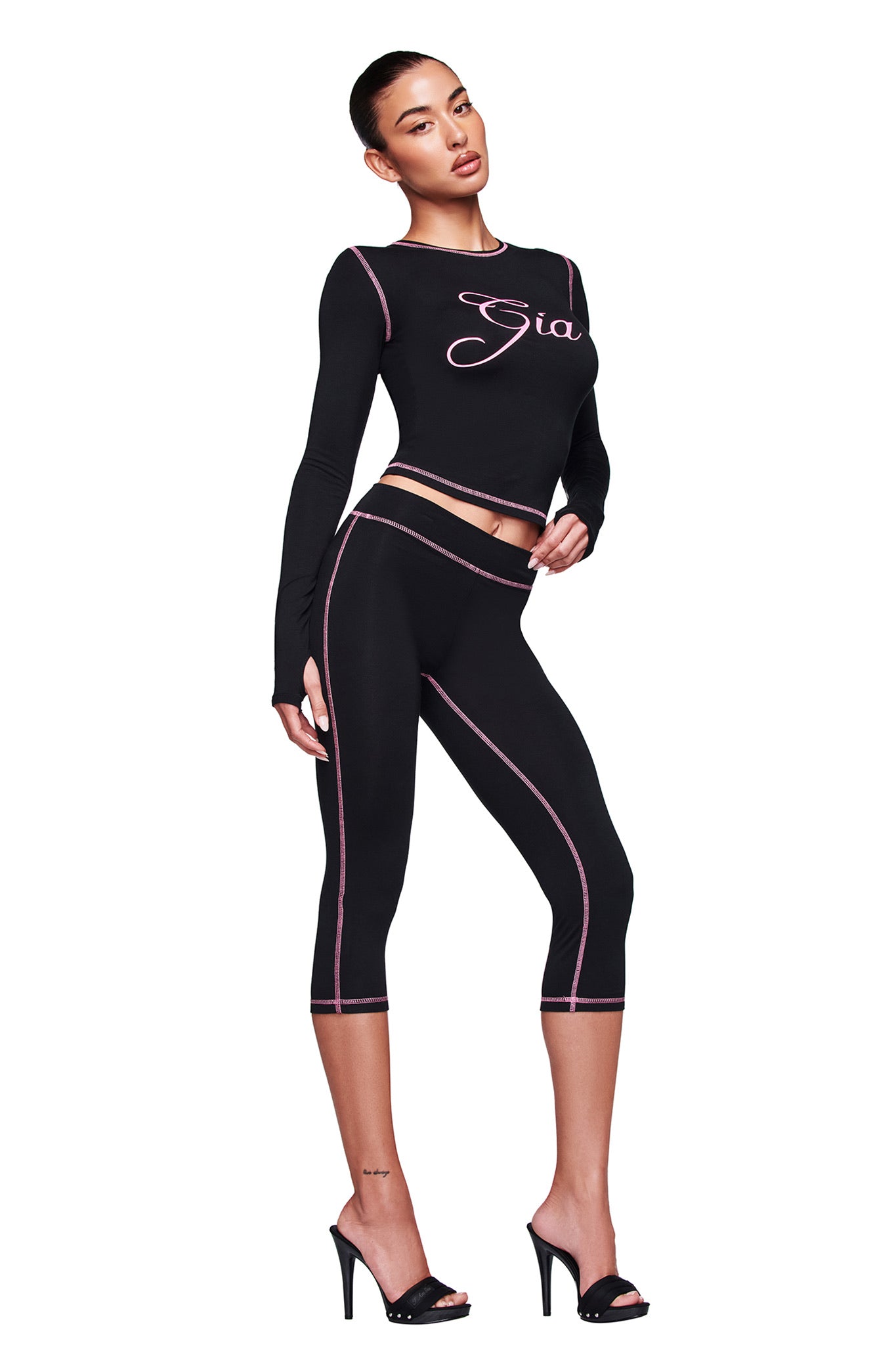 A woman poses in the fitted black BLARE LONG SLEEVE TOP by I.AM.GIA, featuring "Gia" text, with matching black leggings with pink seams and black high-heeled sandals, set against a white background.