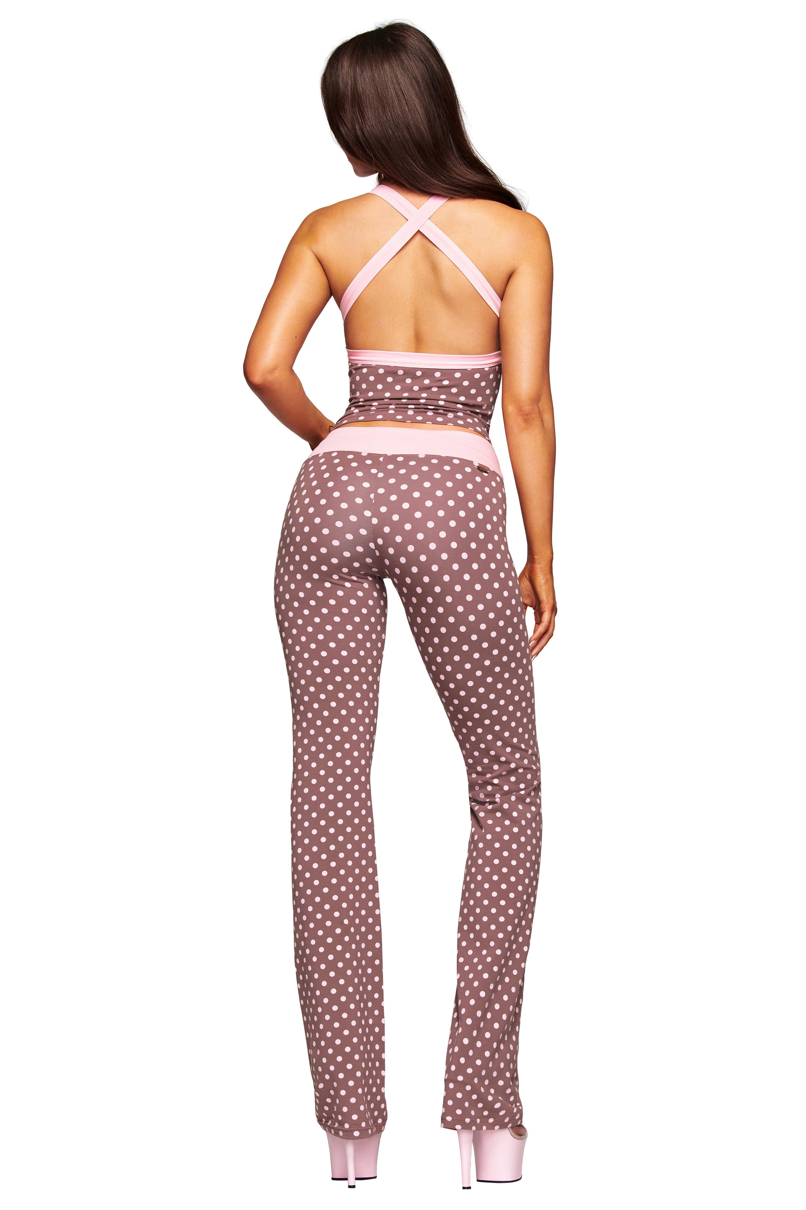 A woman with long hair wears the I.AM.GIA BLARE HALTER STRAP TOP in Pink Chocolate, featuring pink straps and trim, paired with flared white polka dot pants and pink platform heels, facing away from the camera.