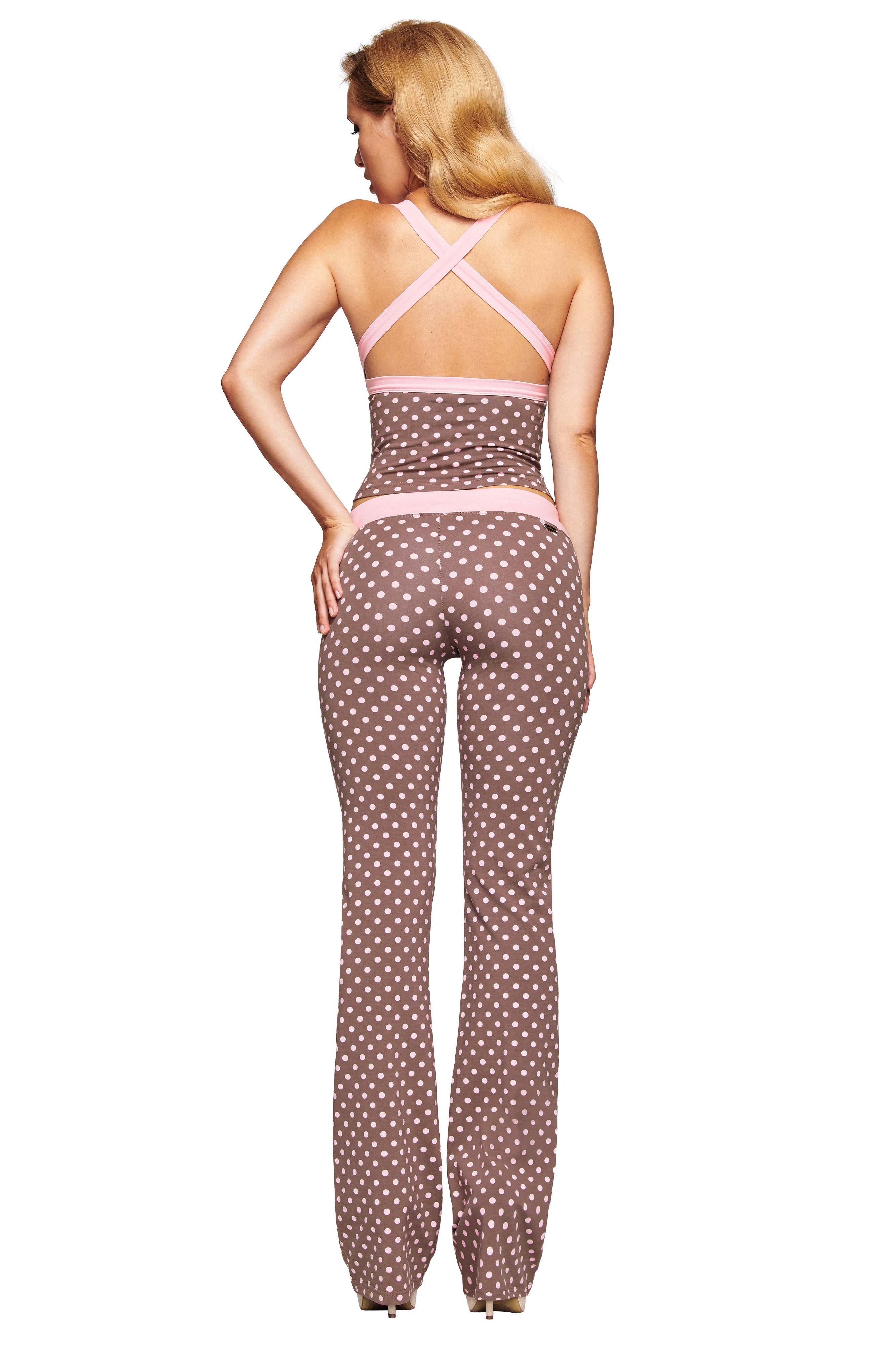 A woman with long blonde hair faces away, wearing the I.AM.GIA BLARE CROSSOVER PANT in Pink Chocolate—a brown polka dot sleeveless jumpsuit with pink trim, flared legs, crossed back straps, and a fitted silhouette.