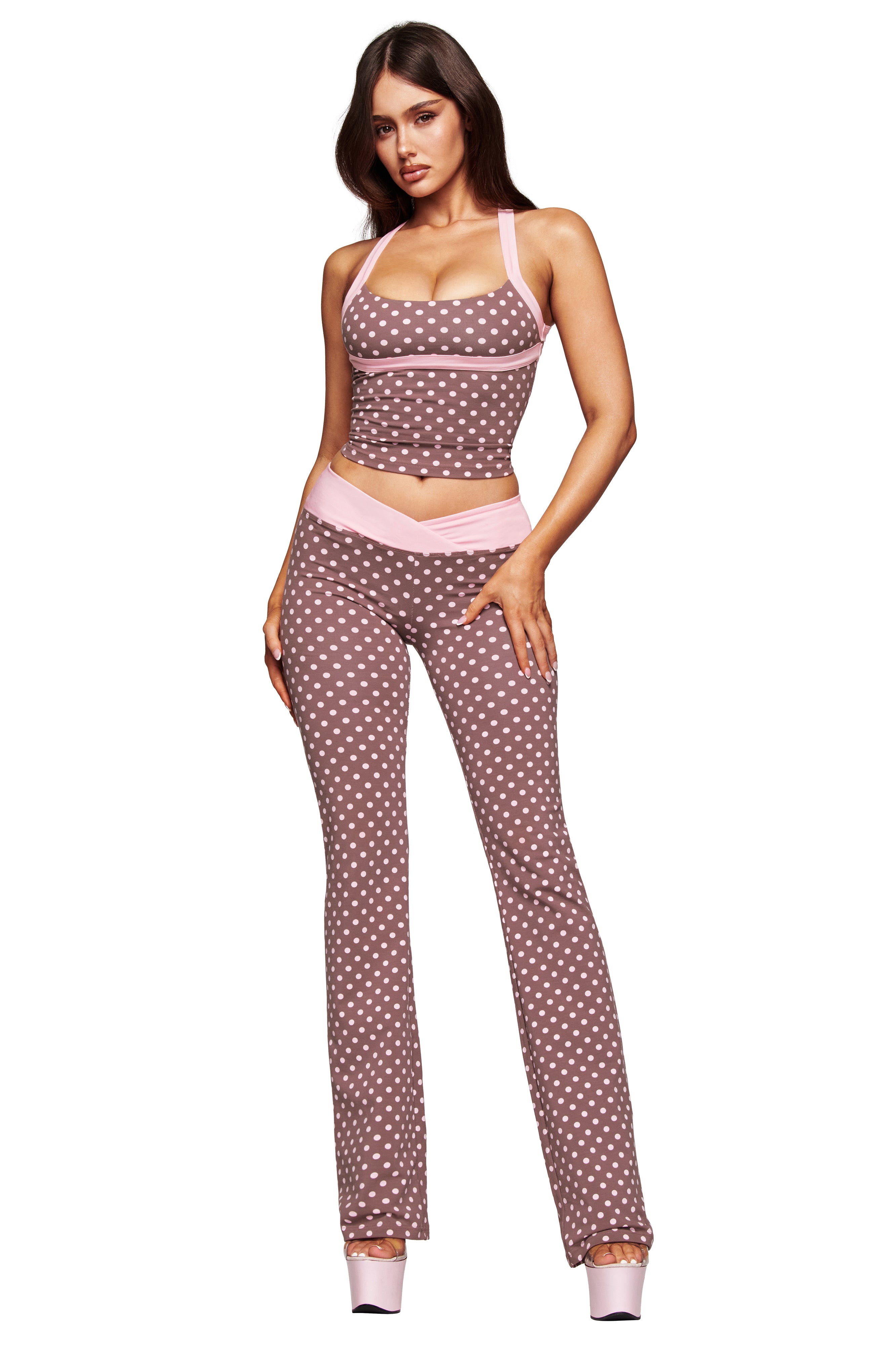 A woman with long brown hair wears the I.AM.GIA BLARE CROSSOVER PANT - PINK CHOCOLATE, featuring a V-shaped waistband and flared legs, paired with a matching sleeveless fitted crop top and platform high heels.