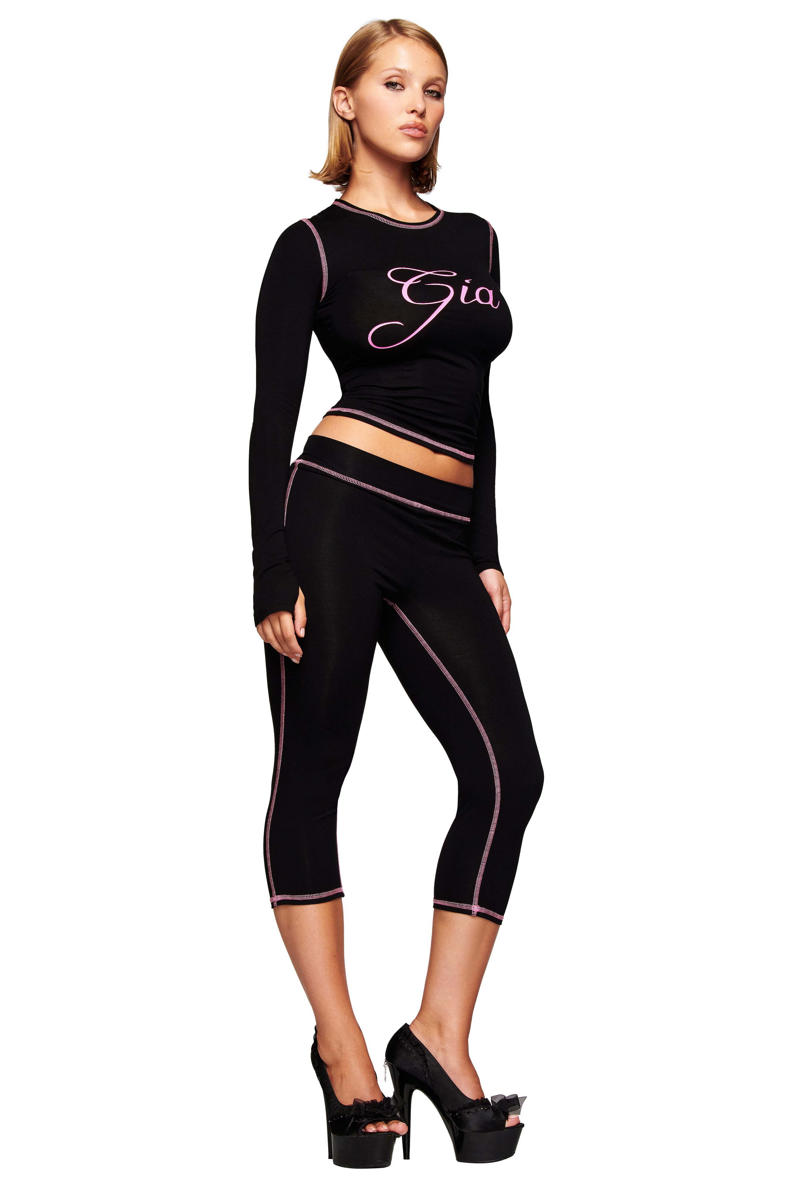 A woman poses confidently in the I.AM.GIA BLARE CAPRI - BLACK with pink stitching, a black I.AM.GIA crop top, and platform heels against a white background.