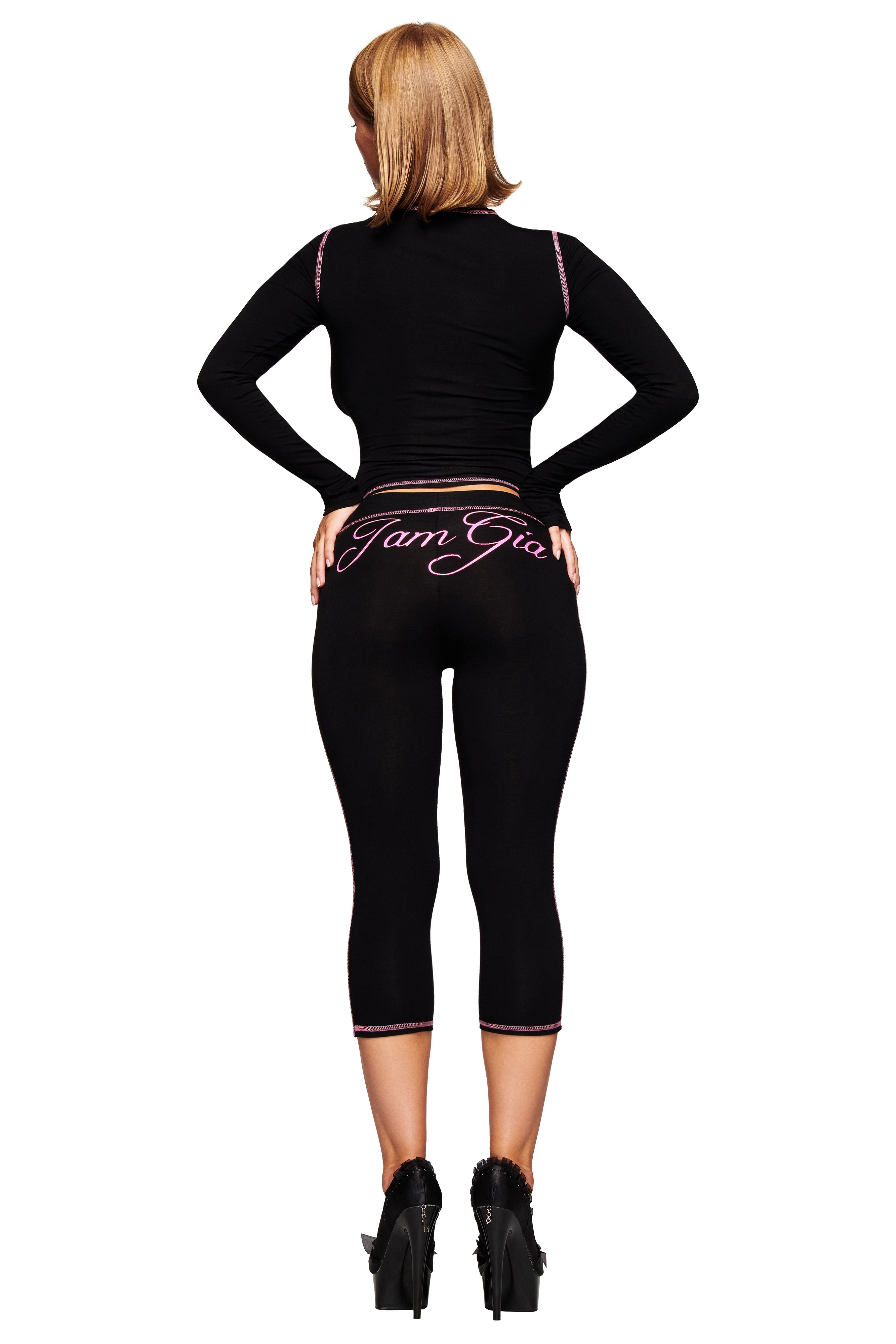 A woman with blond hair stands with hands on hips, facing away, wearing a black long-sleeve crop top and I.AM.GIA BLARE CAPRI - BLACK pants featuring the pink script I.AM.GIA logo on the back.