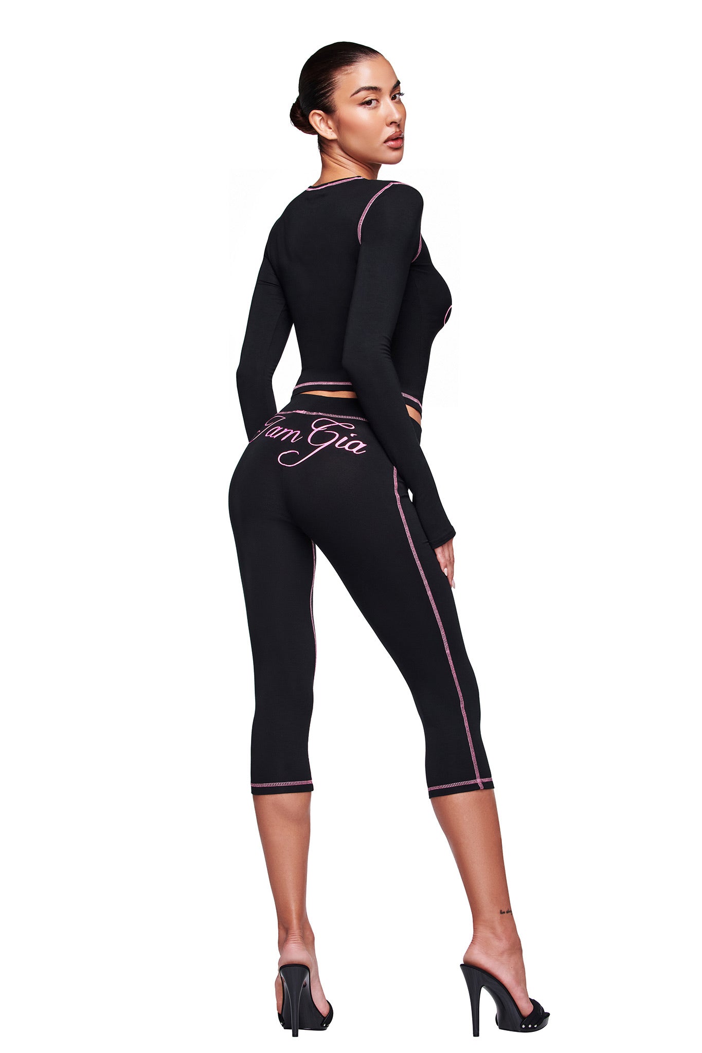 A woman with dark hair in a sleek bun models the I.AM.GIA BLARE CAPRI - BLACK: a black, high-waisted capri pant-style legging with pink trim and script on the back, paired with a matching black long-sleeve crop top and black heeled sandals.