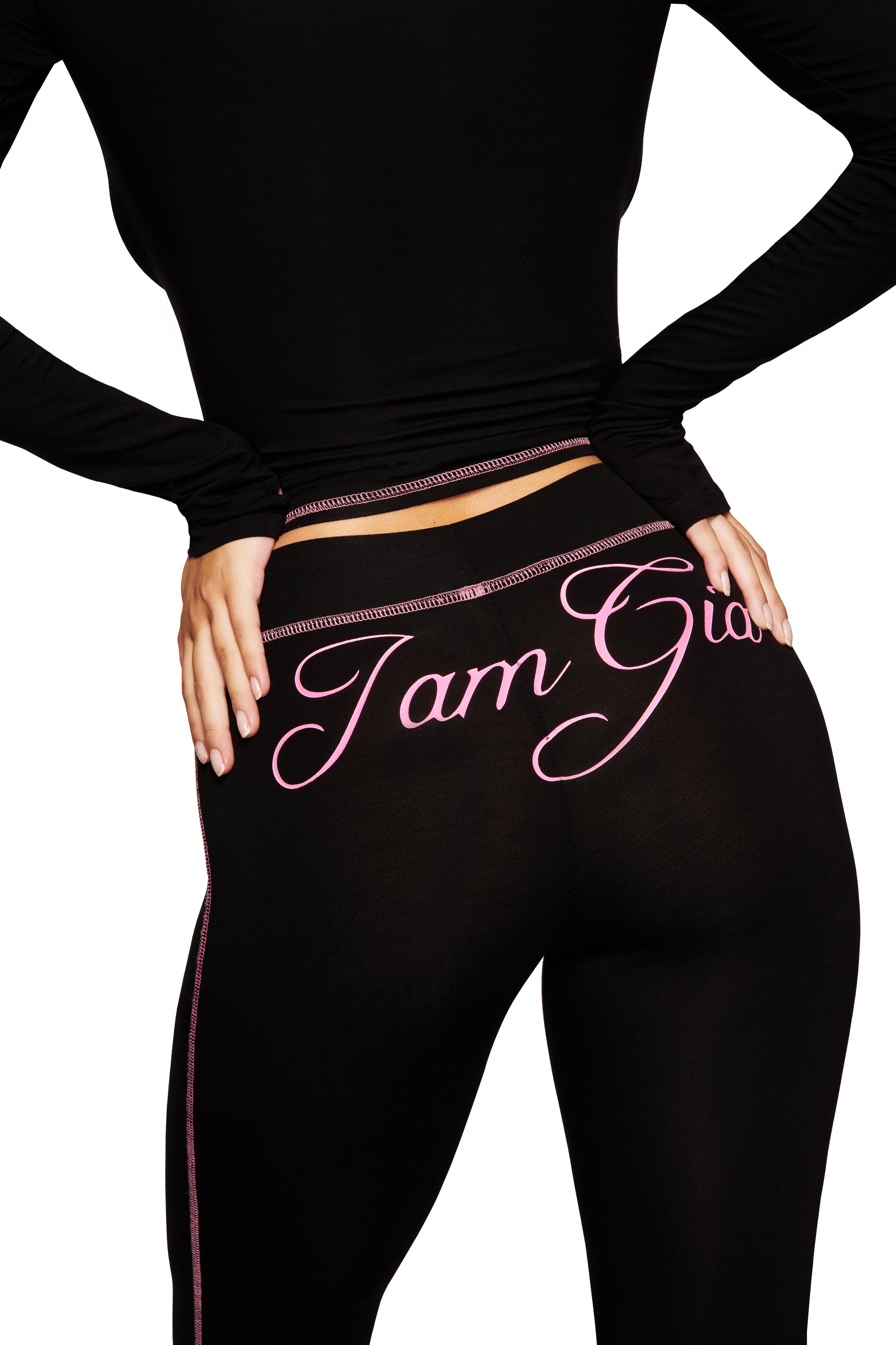 A person stands with hands on hips, wearing I.AM.GIA's BLARE CAPRI in black featuring a pink cursive logo on the back, paired with a matching black long-sleeve top.